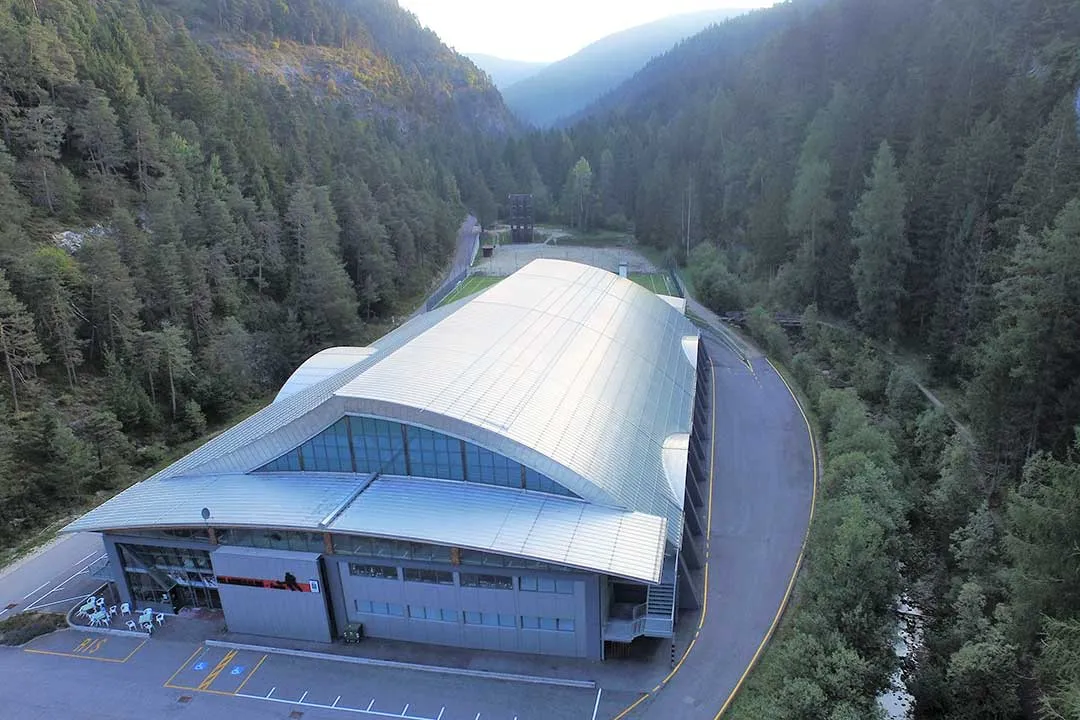 Veduta aerea del palaghiaccio Palasmeraldo con il tetto metallico curvo, l'ingresso e il parcheggio, nella valle boscosa accanto al Lago Smeraldo. – Palasmeraldo (Stadio del ghiaccio)