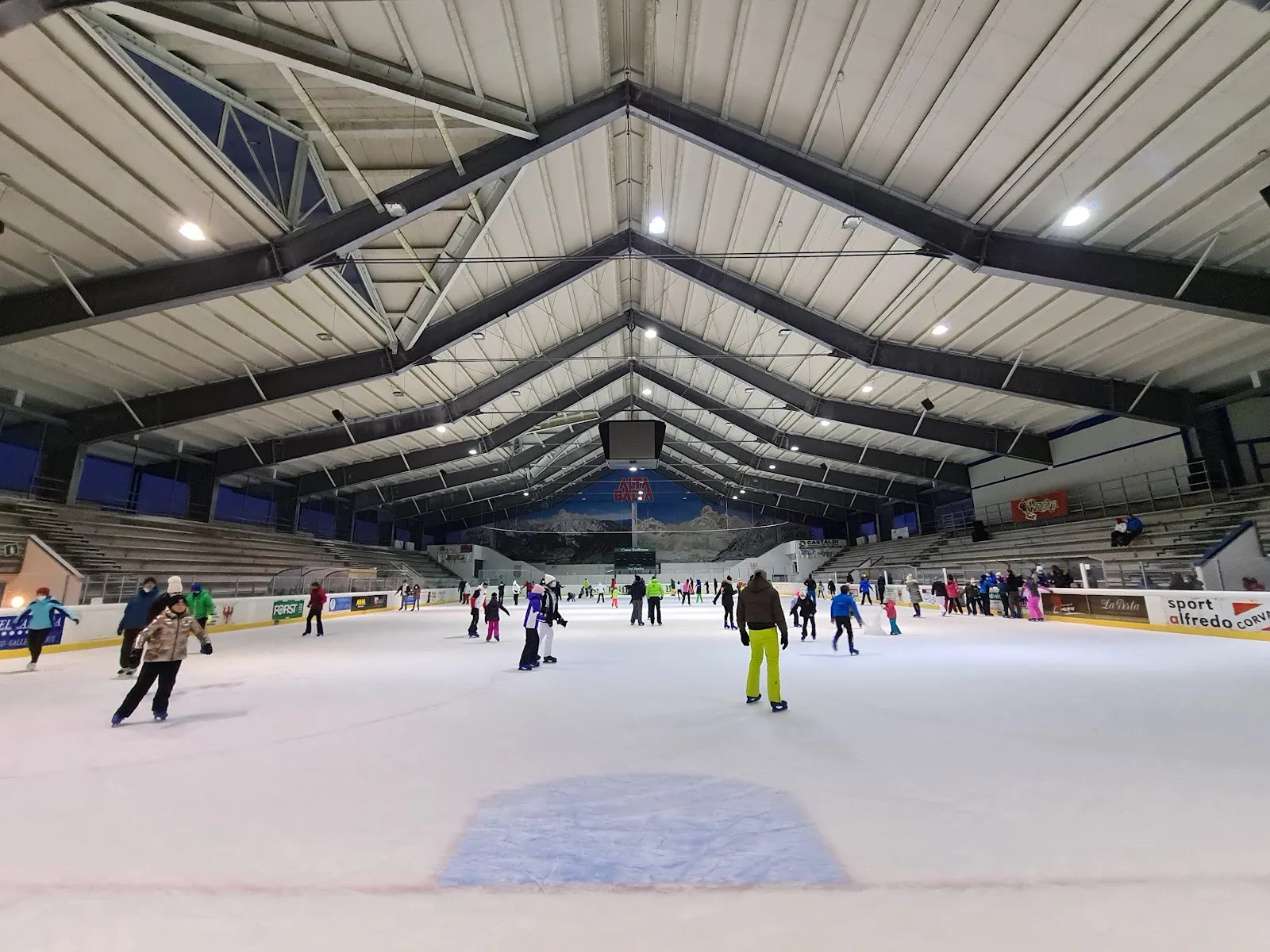 Public skating on the wide ice sheet under the steel-trussed roof, skaters at a distance – Palazzetto del Ghiaccio Corvara (Ice Stadium Corvara)