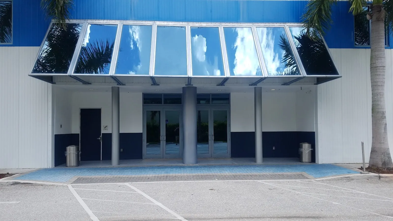 The blue and white glazed entrance of Palm Beach Ice Works with a dark door bay under a glass canopy, seen from the paved forecourt. — Palm Beach Ice Works