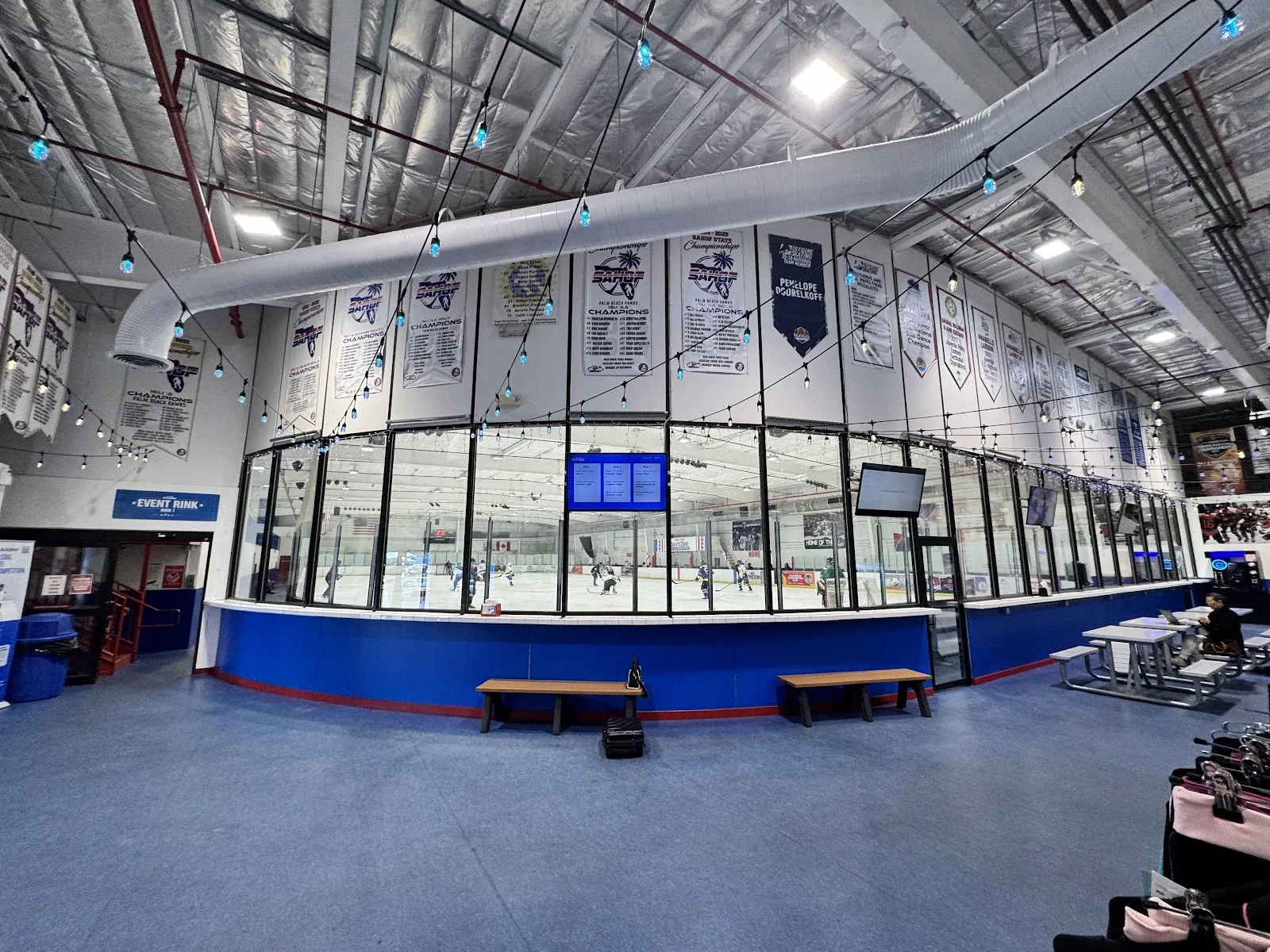 The rink at Palm Beach Skate Zone seen through the curved glass wall of the lobby, with championship banners above and a game far across the ice — Palm Beach Skate Zone (Lake Worth)