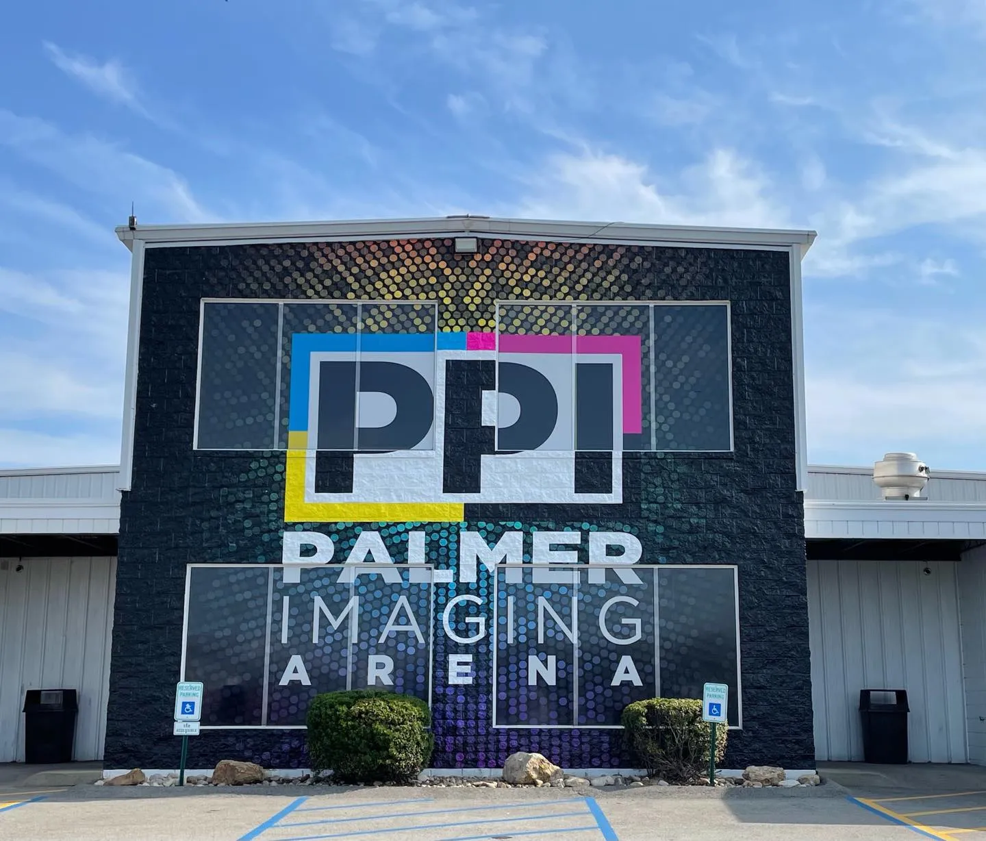 Front of the black-brick building with a large "PPI Palmer Imaging Arena" wall graphic above the entrance, under a clear blue sky — Palmer Imaging Arena