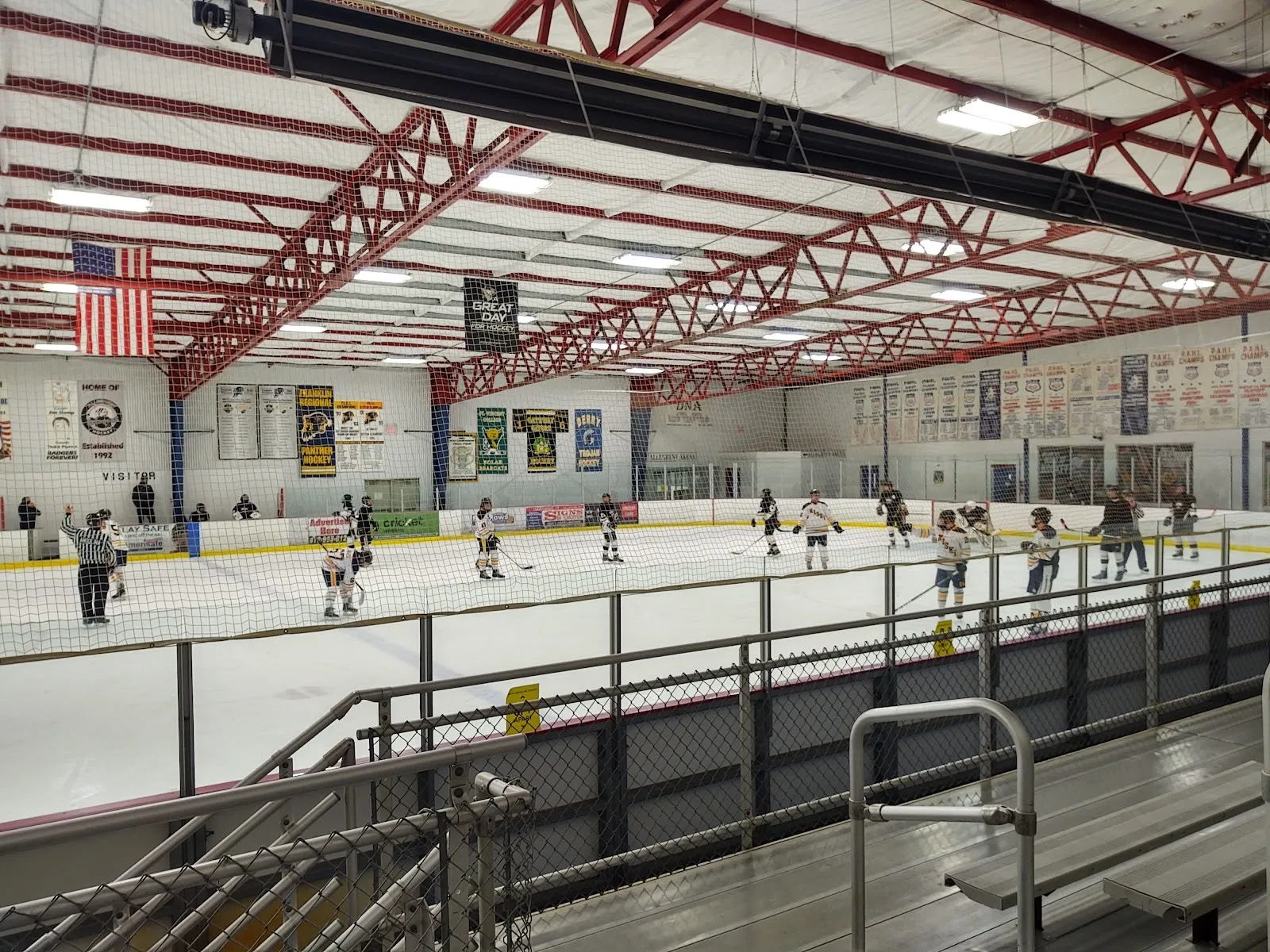 A youth game at Palmer Imaging Arena seen from the bleachers, under red roof trusses with the school hockey banners on the far wall — Palmer Imaging Arena