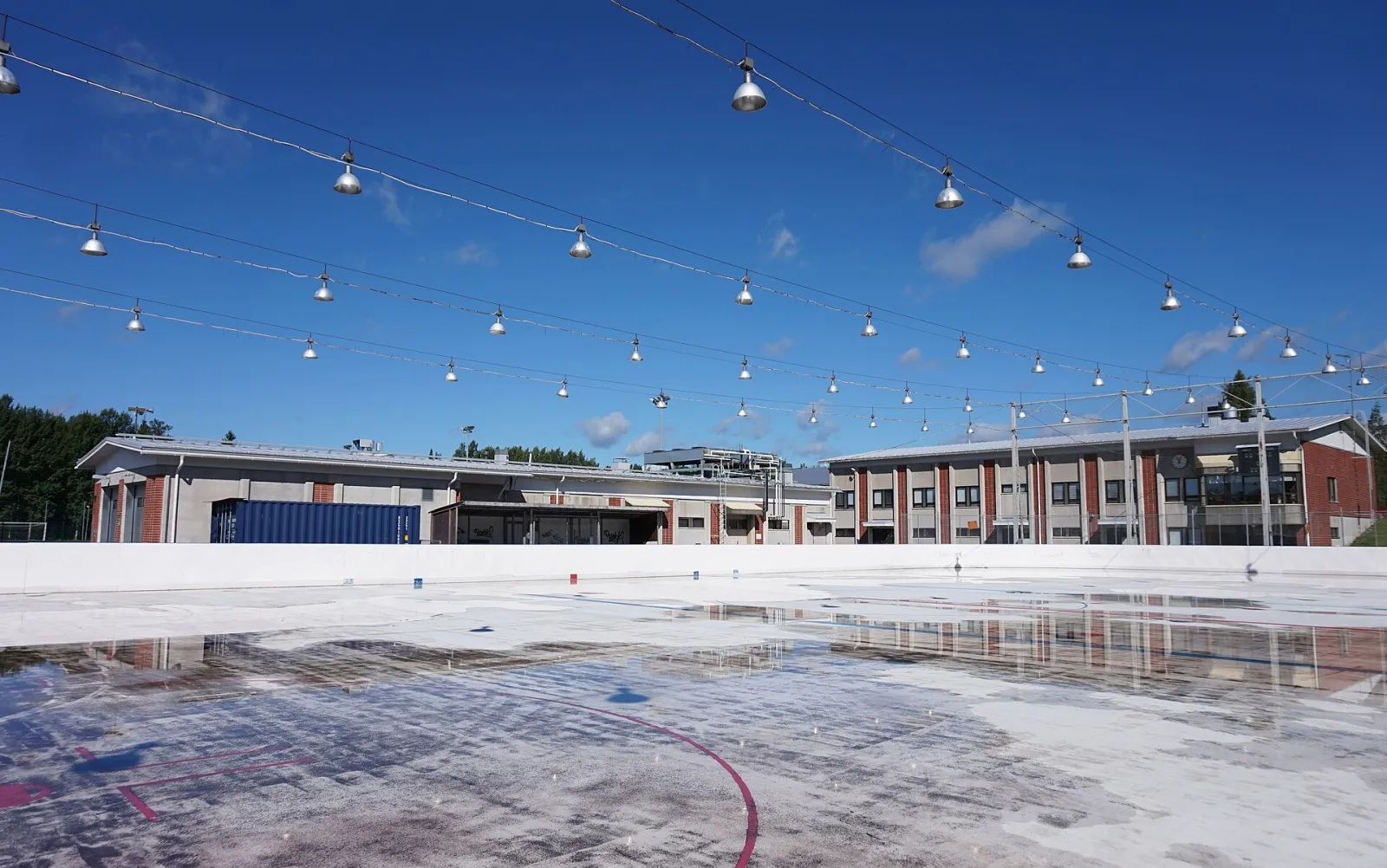 The boarded outdoor ice rink at Palokka sports centre with string lights above it and the buildings behind. — Palokan yhtenäiskoulun luonnonjääkaukalo