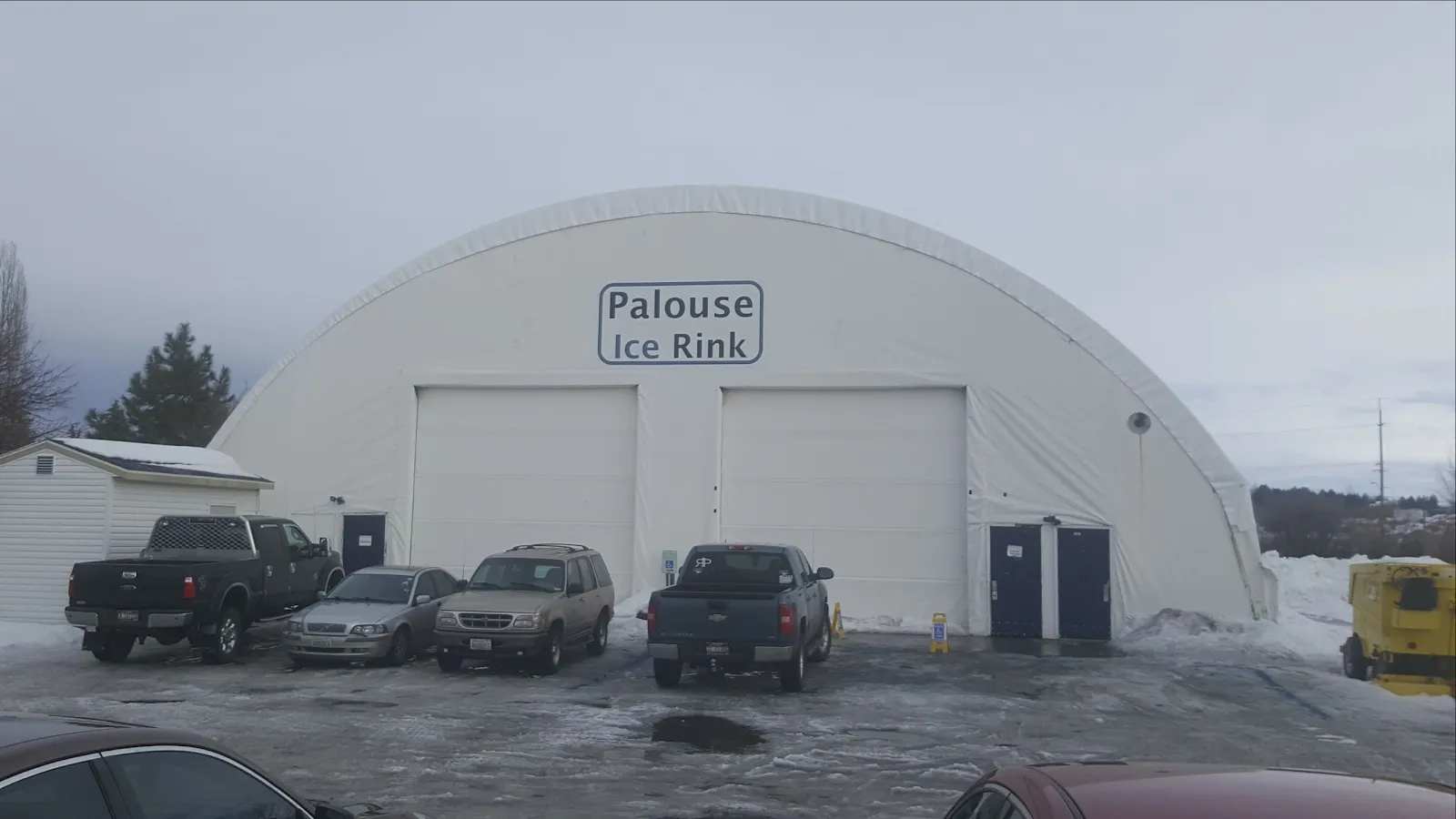The white arched dome housing the Palouse Ice and Roller Rink, a car park with pickup trucks in front under a cloudy sky. — Palouse Ice Rink