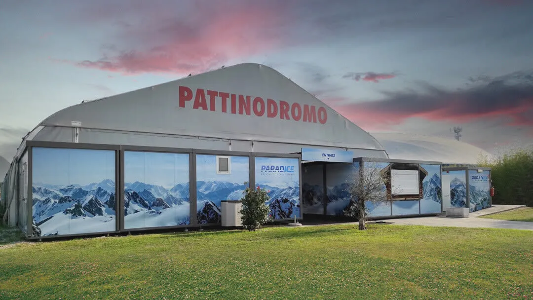 La tensostruttura bianca della pista, con la scritta PATTINODROMO, le vetrate stampate con un panorama di montagna e il nome Paradice sopra l'ingresso, vista oltre il prato. – Paradice Palaghiaccio Montichiari