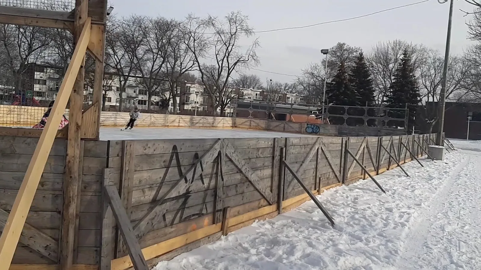 The wooden boards and snow-covered ice of the outdoor rink at Parc De La Bruère with a distant skater and apartment buildings behind — Parc De La Bruère Outdoor Rink