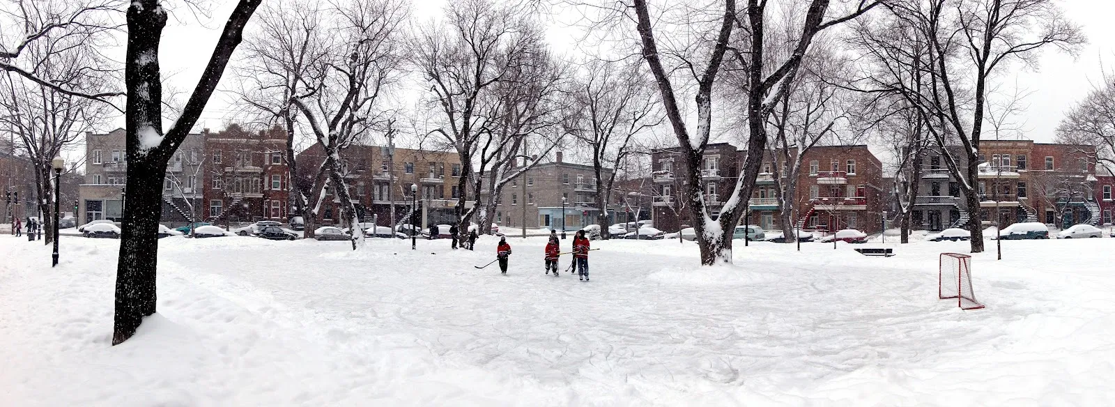 La patinoire extérieure enneigée du parc De Lorimier en hiver, avec des joueurs de hockey et un but au loin et des rangées de triplex montréalais derrière les arbres — Parc De Lorimier Outdoor Rink