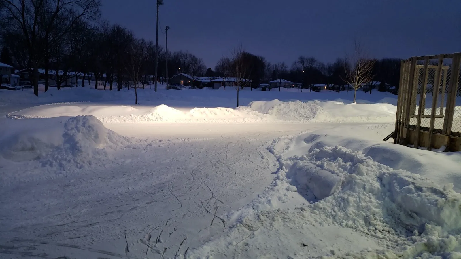 La pista cubierta de nieve de Parc du Père-Frédéric al atardecer, con bancos de nieve alrededor de la superficie de hielo y luces al fondo — Parc du Père-Frédéric Outdoor Rink