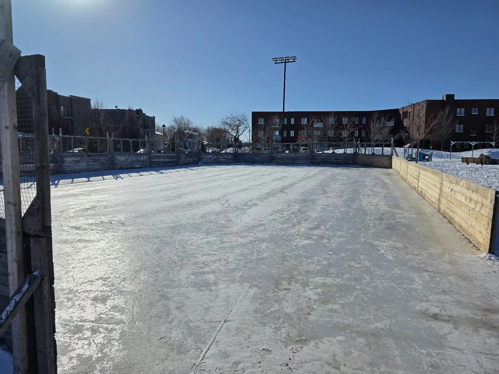 La pista al aire libre de Parc Hans-Selye en un luminoso día de invierno, una pista de hielo congelada rodeada de vallas de madera con edificios de departamentos de ladrillo detrás — Parc Hans-Selye Outdoor Rink