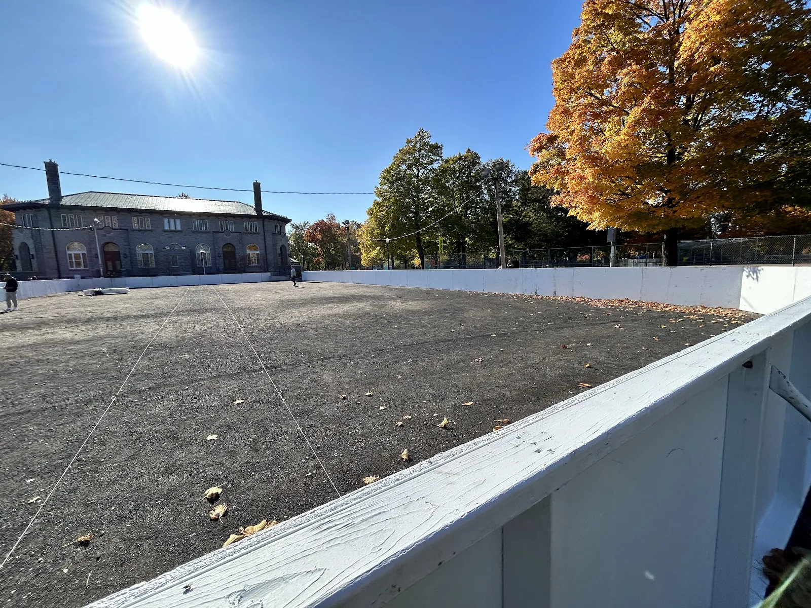 A rink with white boards at Parc La Fontaine in autumn, bare surface, with the park building behind, no people in view — Parc La Fontaine Outdoor Rinks