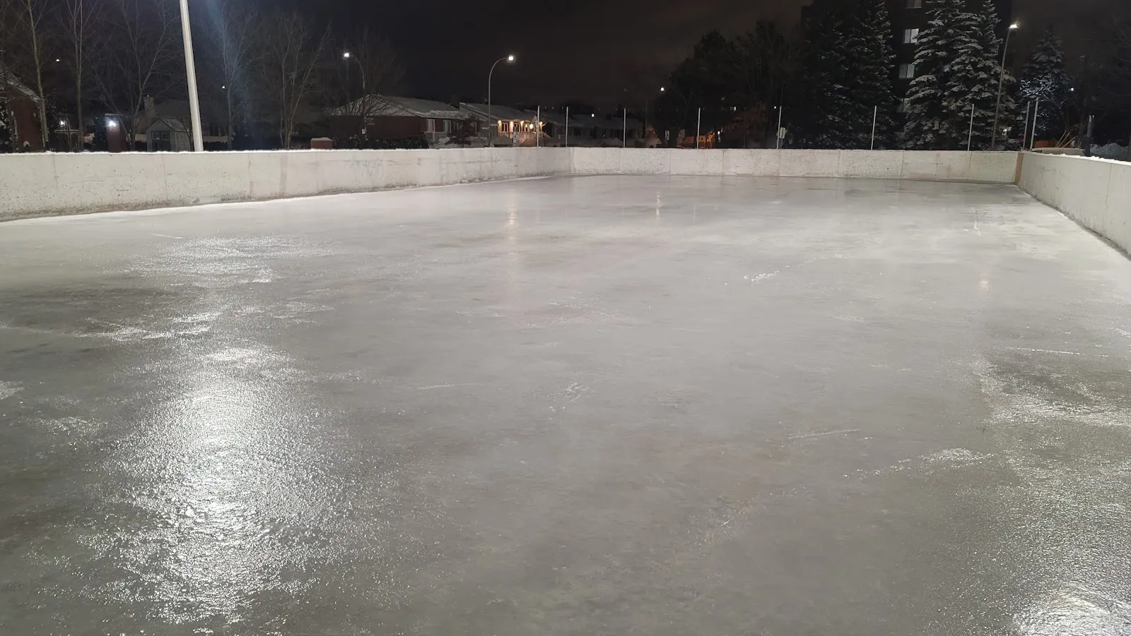 Freshly flooded ice on the boarded outdoor rink at Parc Lucie-Bruneau at night, a smooth surface under park lights with no people in view — Parc Lucie-Bruneau Outdoor Rinks