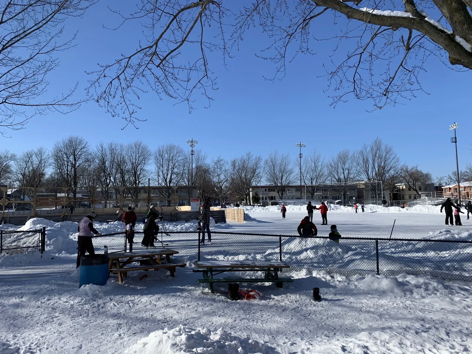 Skaters and children on the snow-banked outdoor rink at Parc Saint-Victor behind a chain-link fence and picnic tables, with bare trees and rink floodlights under a clear blue winter sky. — Parc Saint-Victor Outdoor Rinks