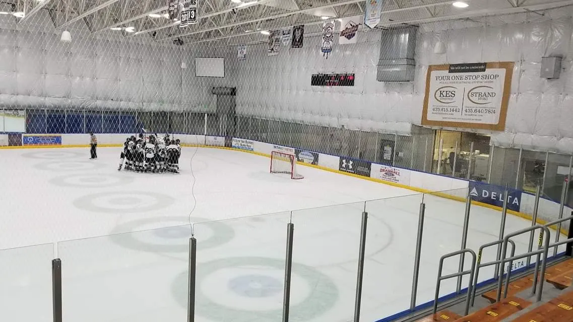 Curling circles on the white sheet beyond glass and orange boards, a team gathered at one end and a scoreboard and banners on the far wall. — Park City Ice Arena