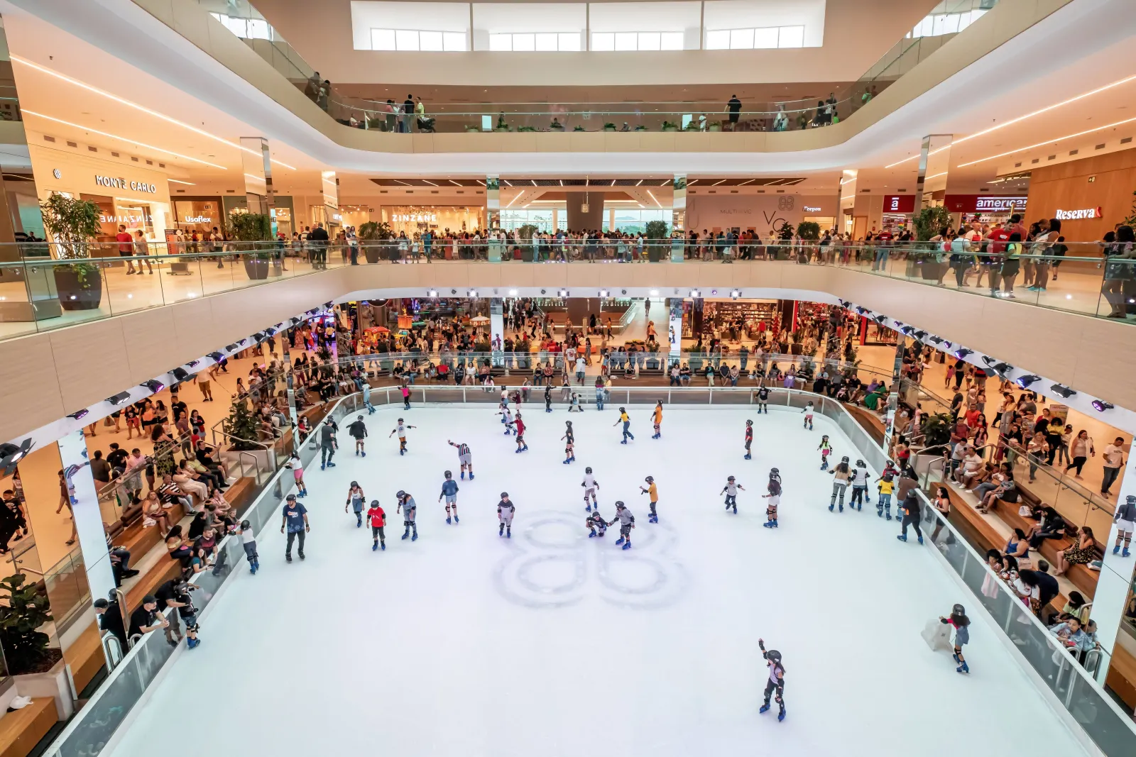 The mall's permanent ice rink seen from the upper floors, with skaters spread across the ice under the atrium and spectators on the benches along the rail — ParkJacarepaguá Ice Rink