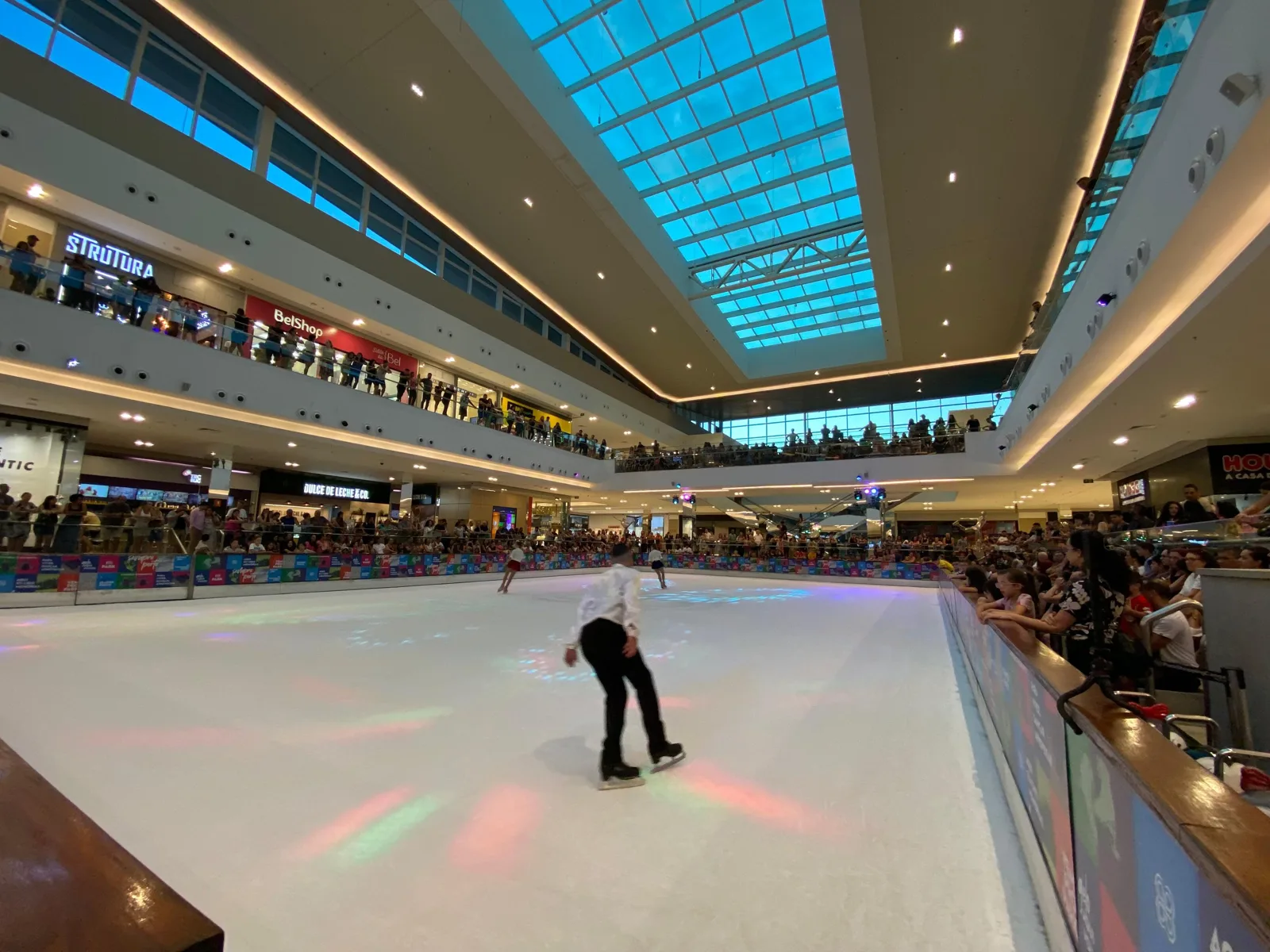 Um patinador atravessa o gelo branco da pista de 450 metros quadrados, com espectadores junto ao guarda-corpo e o átrio com claraboia do shopping ao fundo. — ParkShopping Canoas Ice Rink