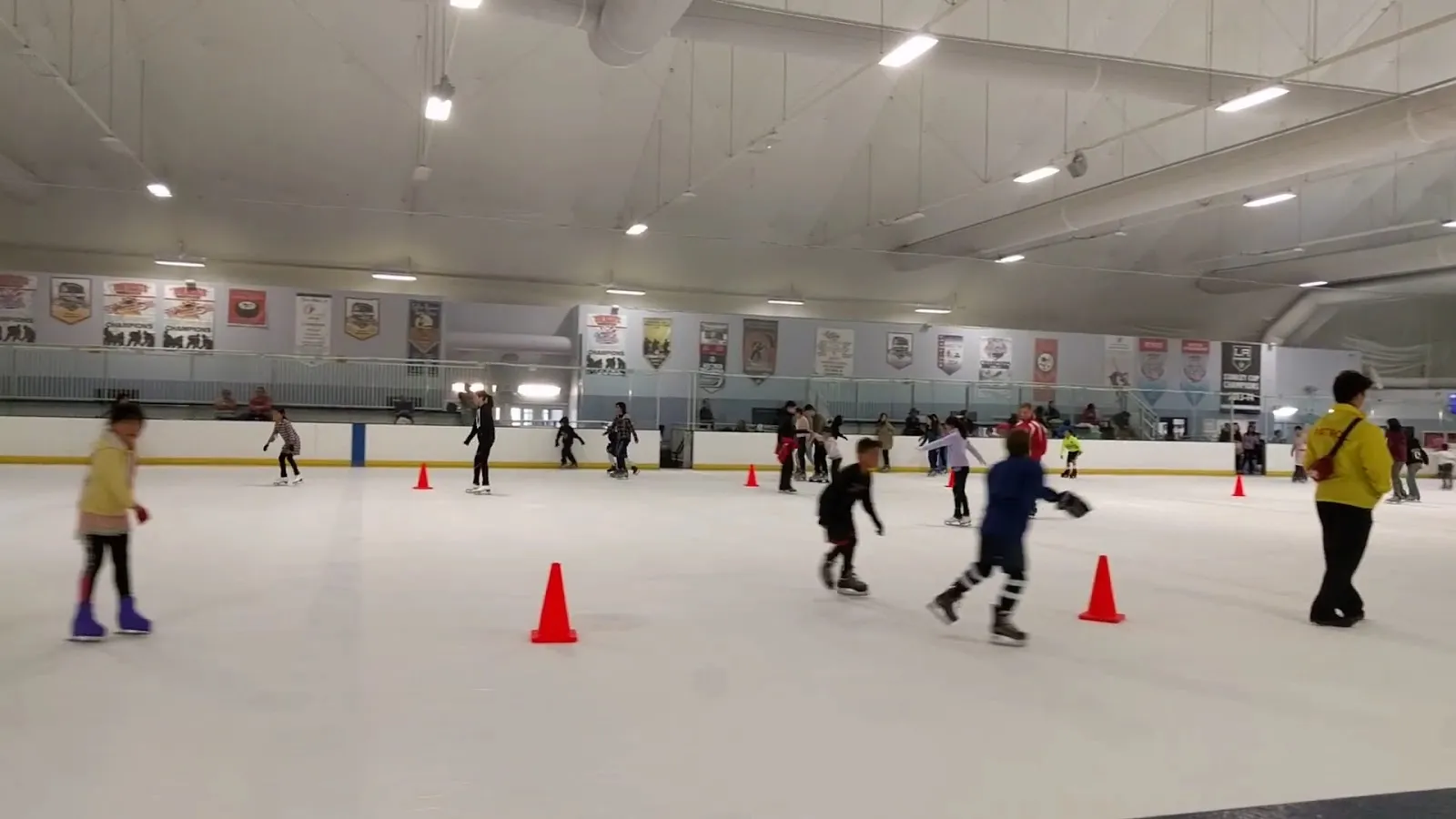 Una sesión pública en una pista amplia, con patinadores repartidos entre conos naranjas bajo un techo claro con banderines en la pared del fondo. — Pasadena Ice Skating Center
