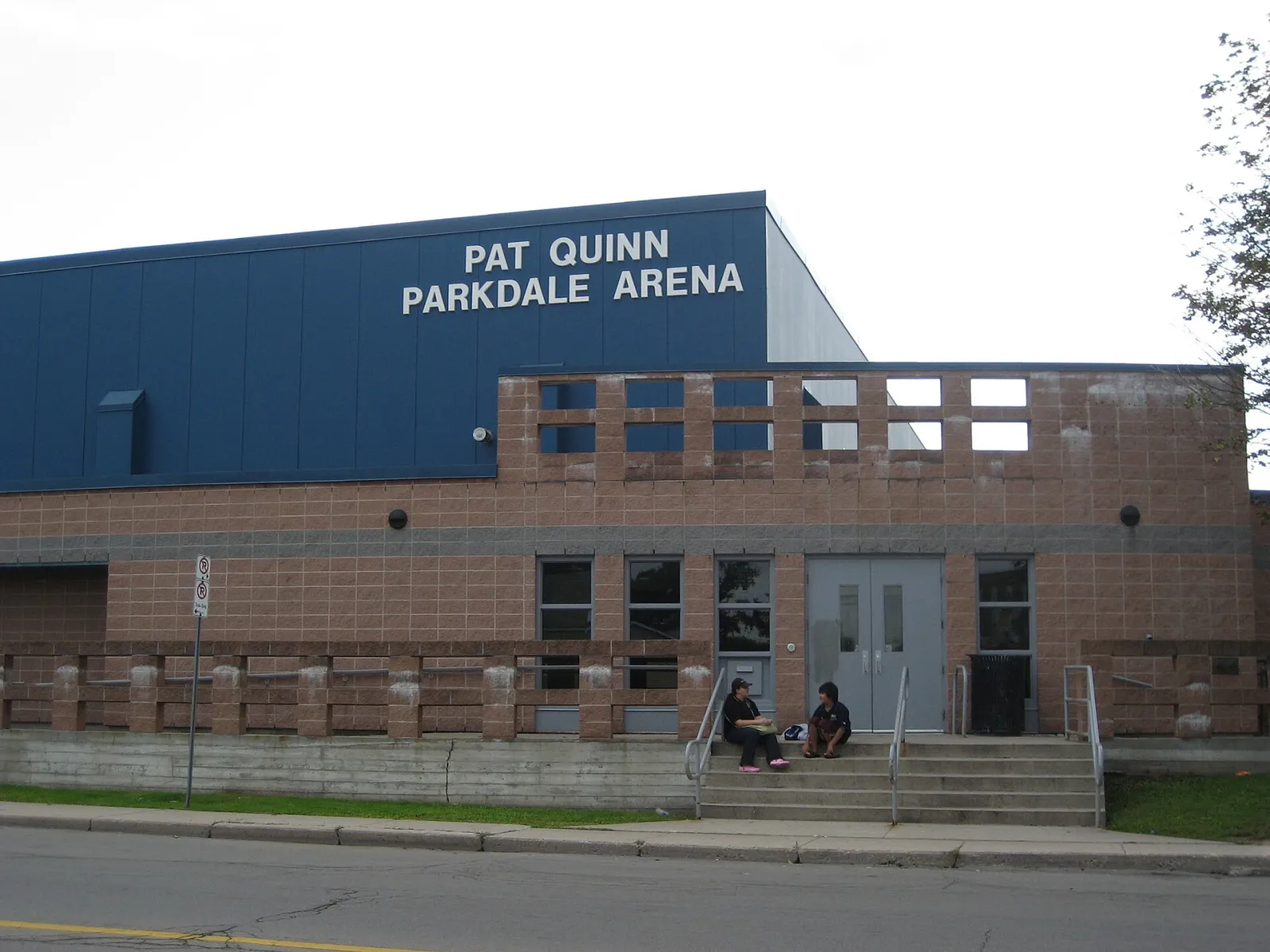 Muro superior azul con la leyenda "Pat Quinn Parkdale Arena" sobre una entrada de ladrillo con escalones que bajan a la banqueta. — Pat Quinn Parkdale Arena