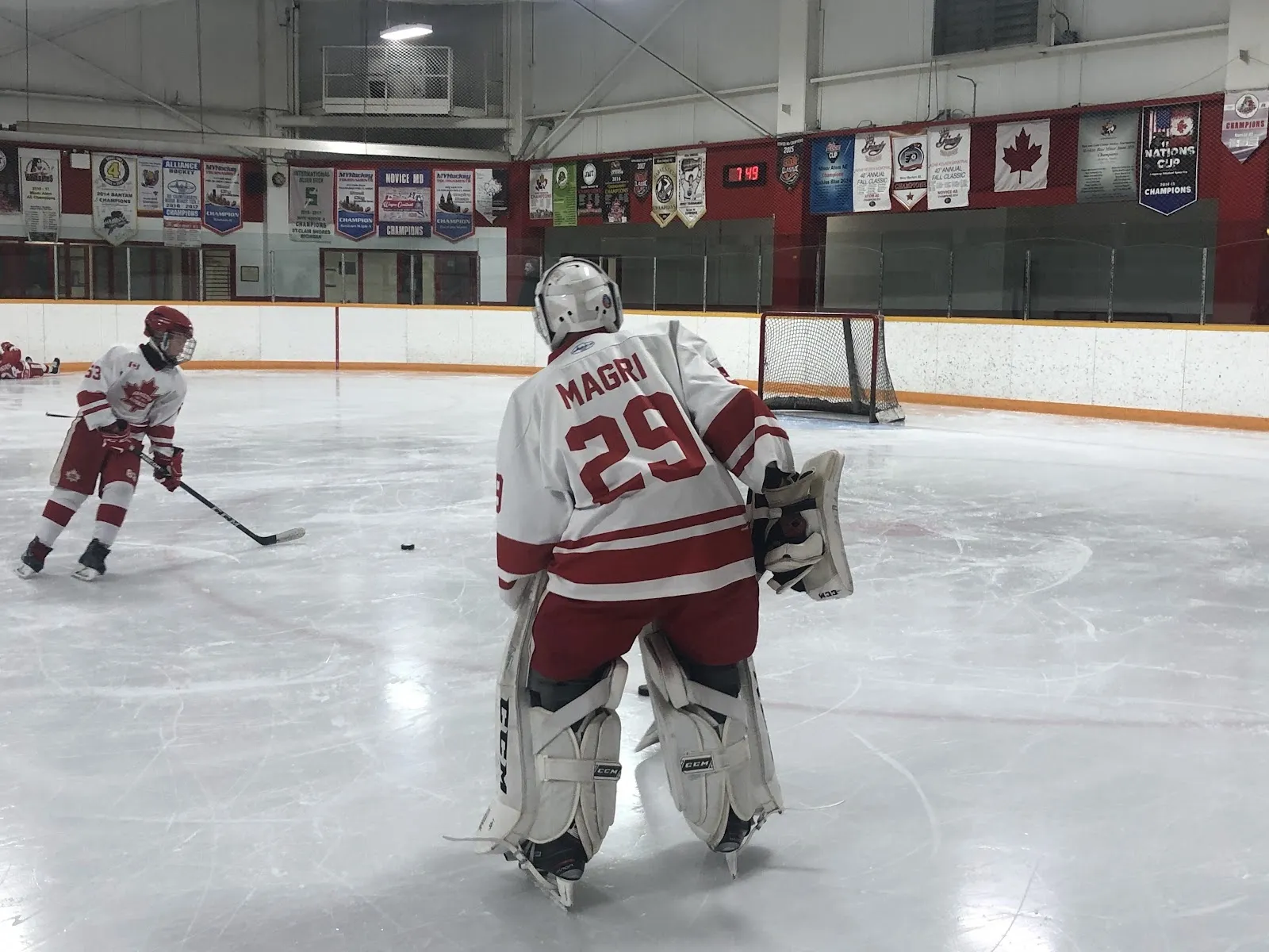Un portero visto de espaldas con jersey rojo y blanco en la portería, con jugadores repartidos por el hielo más allá, y vallas con ribete rojo y banderines detrás. — Pat Quinn Parkdale Arena