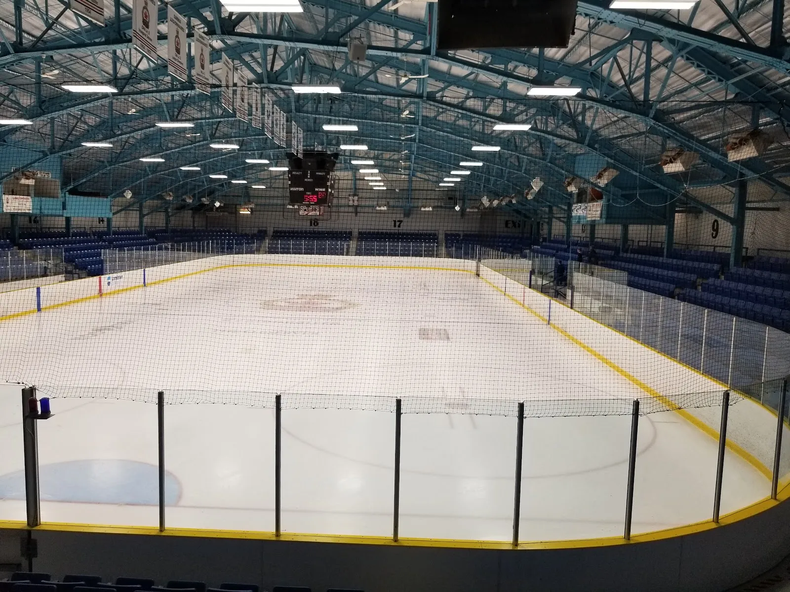 El hielo vacío bajo las armaduras arqueadas color verde azulado del techo, rodeado de asientos azules y red de protección. — Pat Stapleton Arena