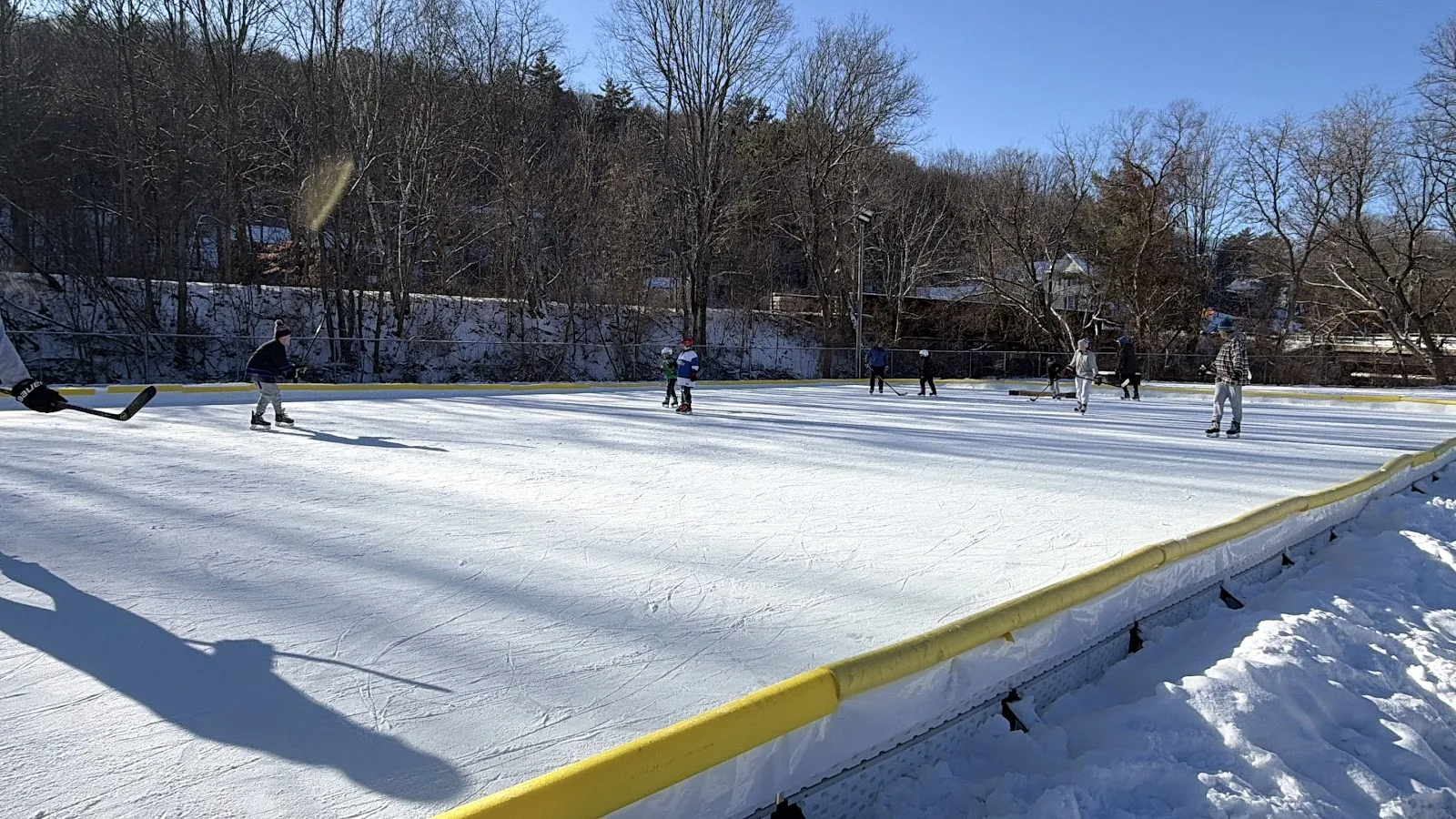 Skaters seen from a distance on the snow-banked, yellow-edged Pat Walsh rink in winter sun — Pat Walsh Ice Rinks