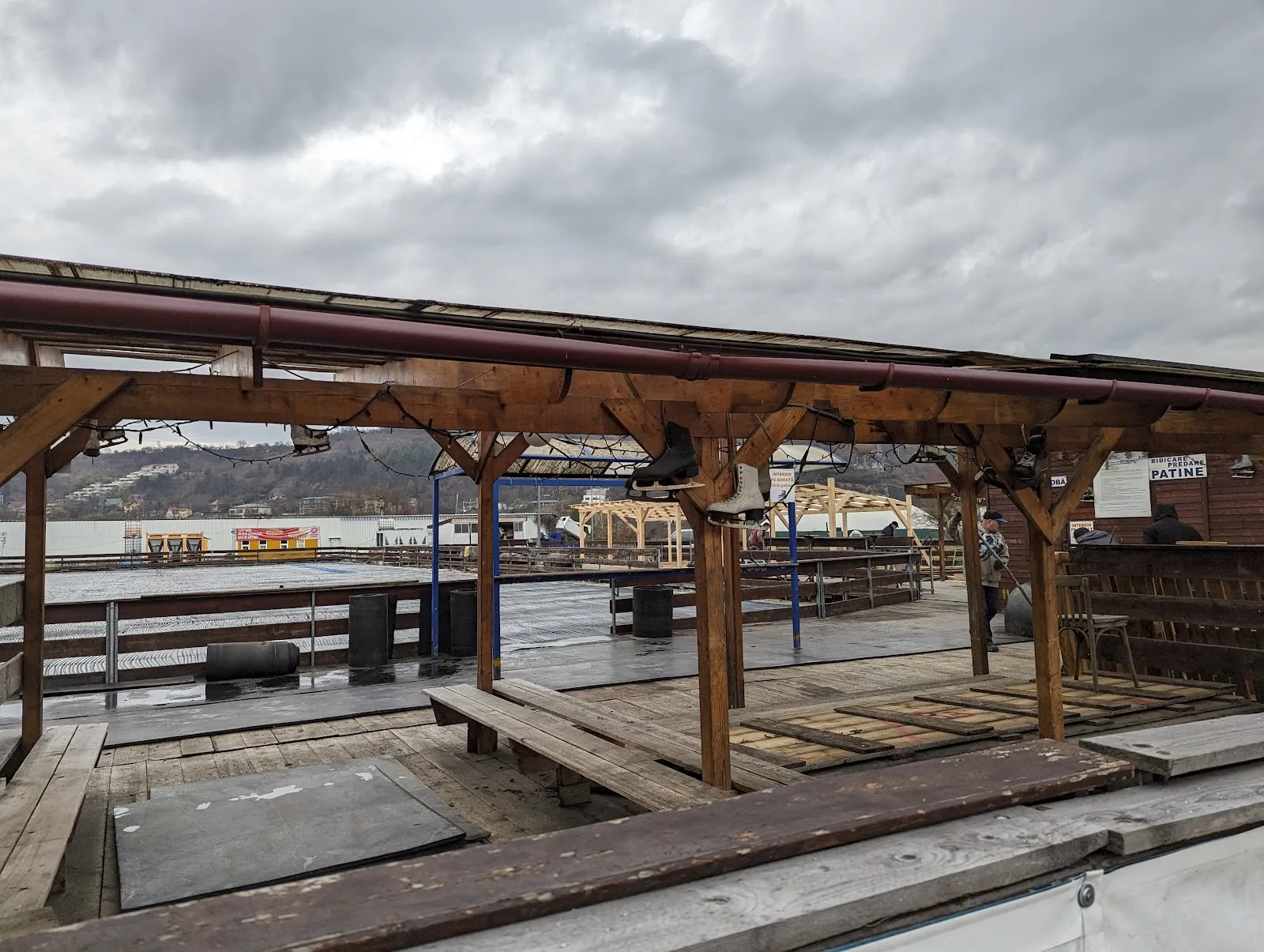 The rink's timber shelter and terrace in the off-season beside the hypermarket car park, with the hills of Cluj beyond. — Patinoar Fiesta Sport