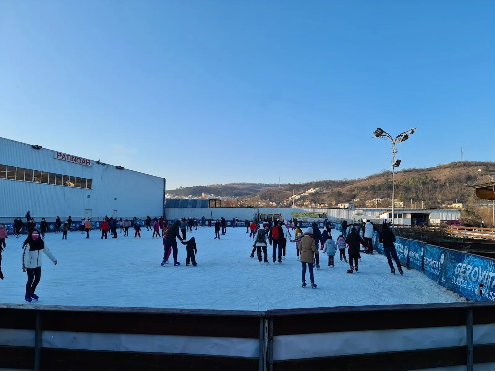 The open-air artificial ice on a sunny winter day, with skaters spread across the sheet inside the dark barrier and the hillside beyond. — Patinoar Fiesta Sport