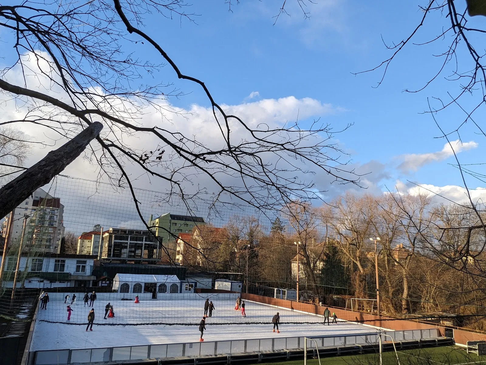 The rink inside its net enclosure with skaters on the ice and the surrounding park trees and apartment blocks behind it. — Patinoar Sibiu