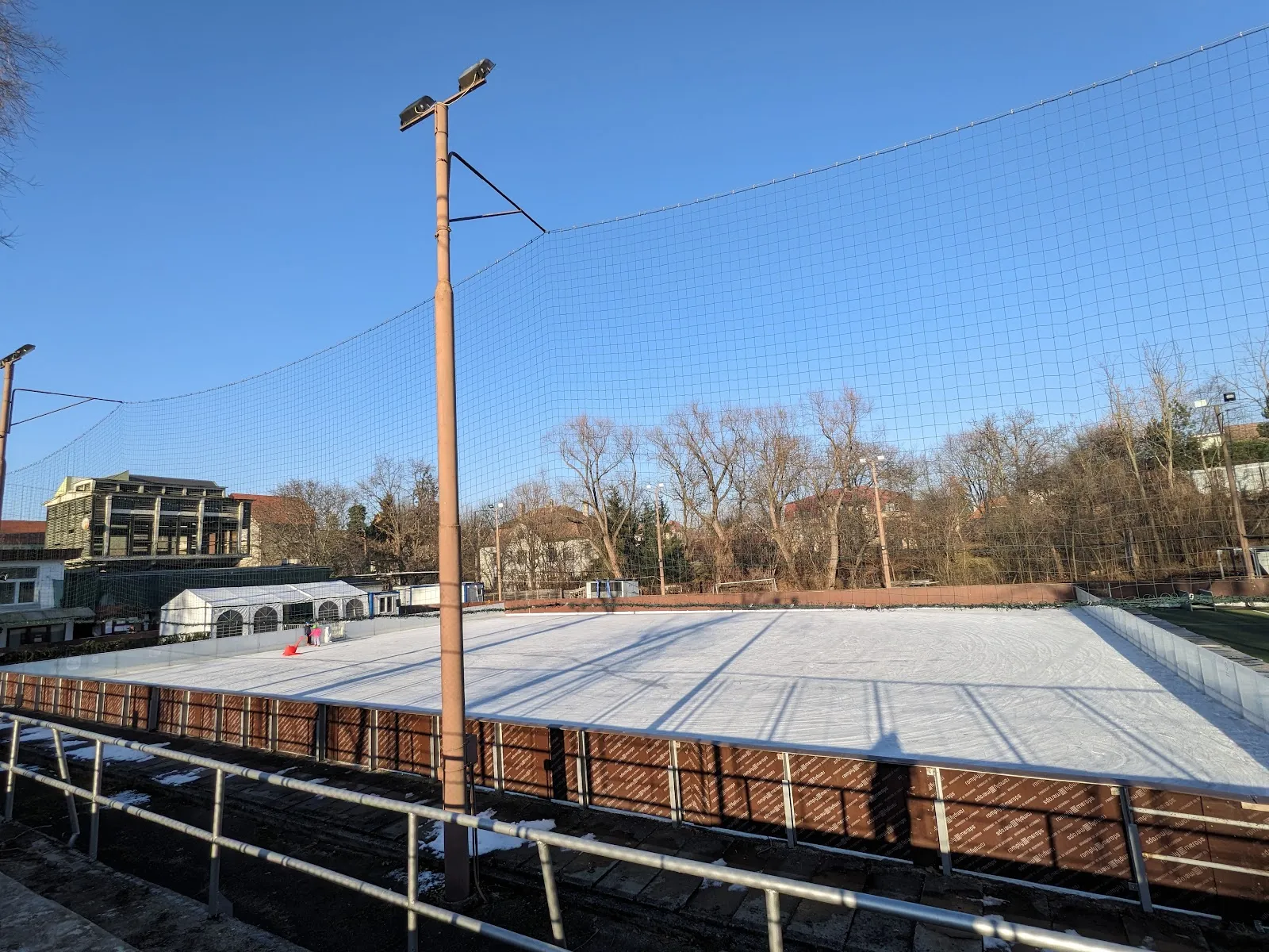 The open-air ice in winter sun, seen from the stand over the wooden barrier, with a floodlight pole and the trees of the park behind. — Patinoar Sibiu