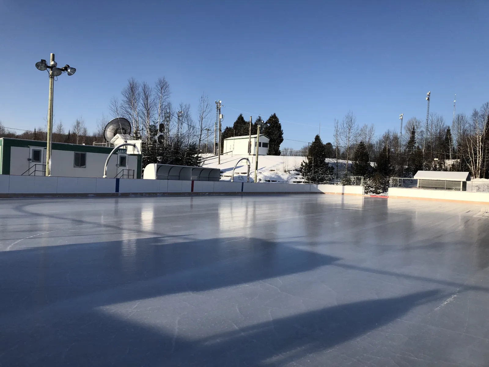 The empty boarded rink at the parc-école in sunshine, with floodlight masts and the service trailer behind the boards. — Patinoire à bandes du parc-école (Sainte-Marguerite-du-Lac-Masson)