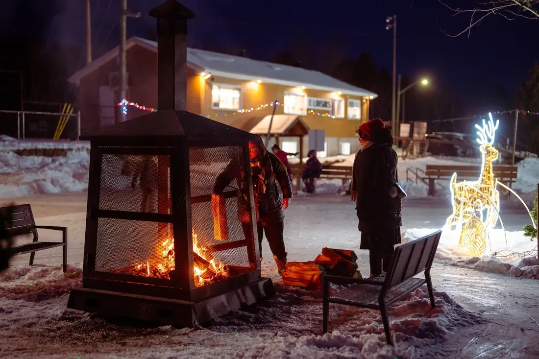 The Saint-Damien rink area at night, with a glowing fire pit, festive reindeer lights, benches and the lit service building beside the ice. — Patinoire, anneau de glace et glissade de Saint-Damien