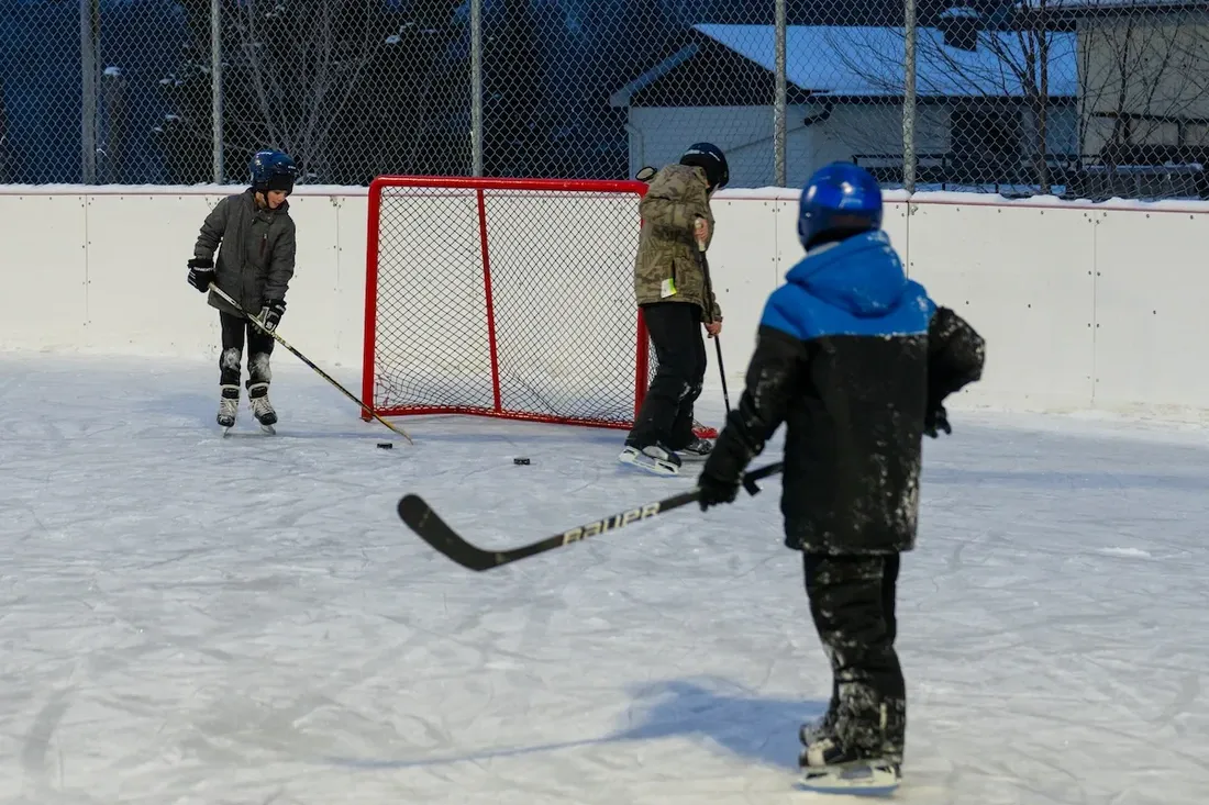 Children playing hockey in front of a goal on the boarded outdoor rink at Saint-Damien, with a building behind the boards. — Patinoire, anneau de glace et glissade de Saint-Damien