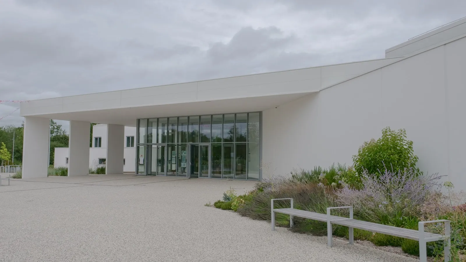 Le bâtiment d’entrée blanc, vitré et à toit plat de la patinoire, avec un parvis gravillonné et des massifs plantés. — Patinoire Arago