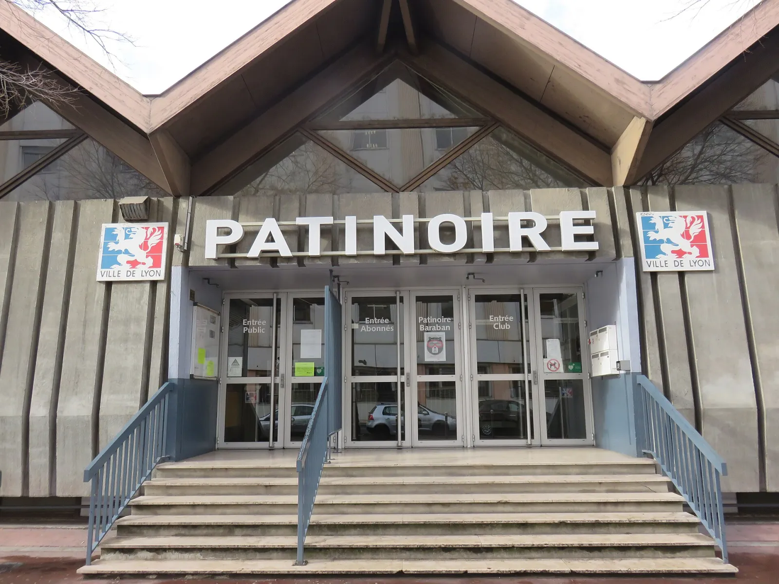 L’entrée de la patinoire Baraban : PATINOIRE en lettres blanches au-dessus des portes vitrées, avec les armes de la Ville de Lyon de part et d’autre et un escalier — Patinoire Baraban