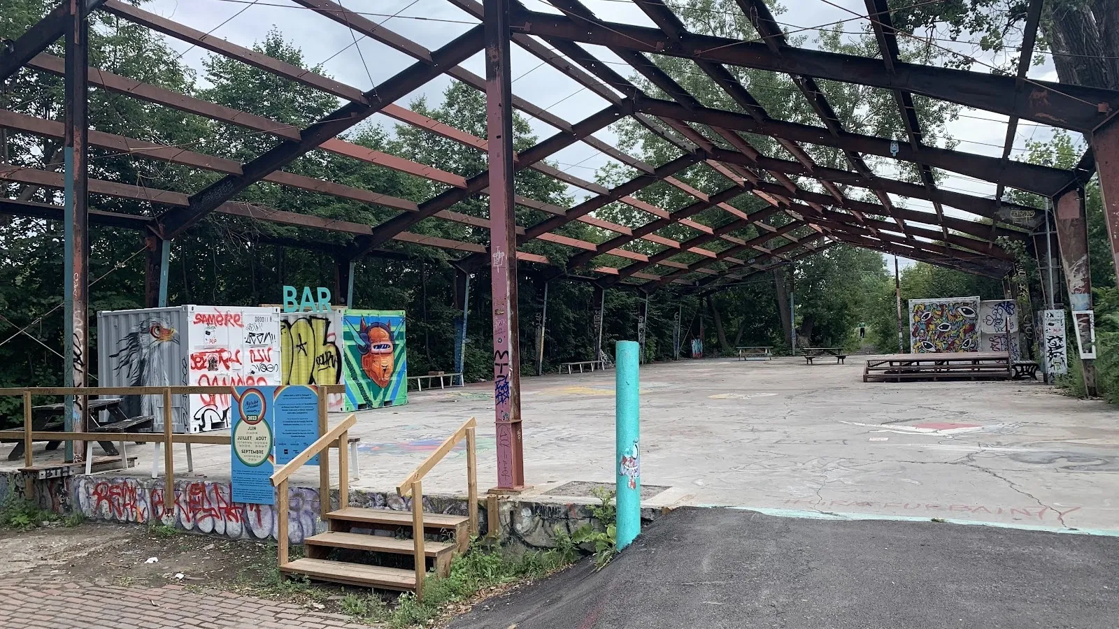 La charpente d’acier du toit de la patinoire au-dessus d’une dalle de béton nue en été, avec des conteneurs maritimes peints, des tables de pique-nique et des graffitis sur le pourtour. — Patinoire Bernard Est