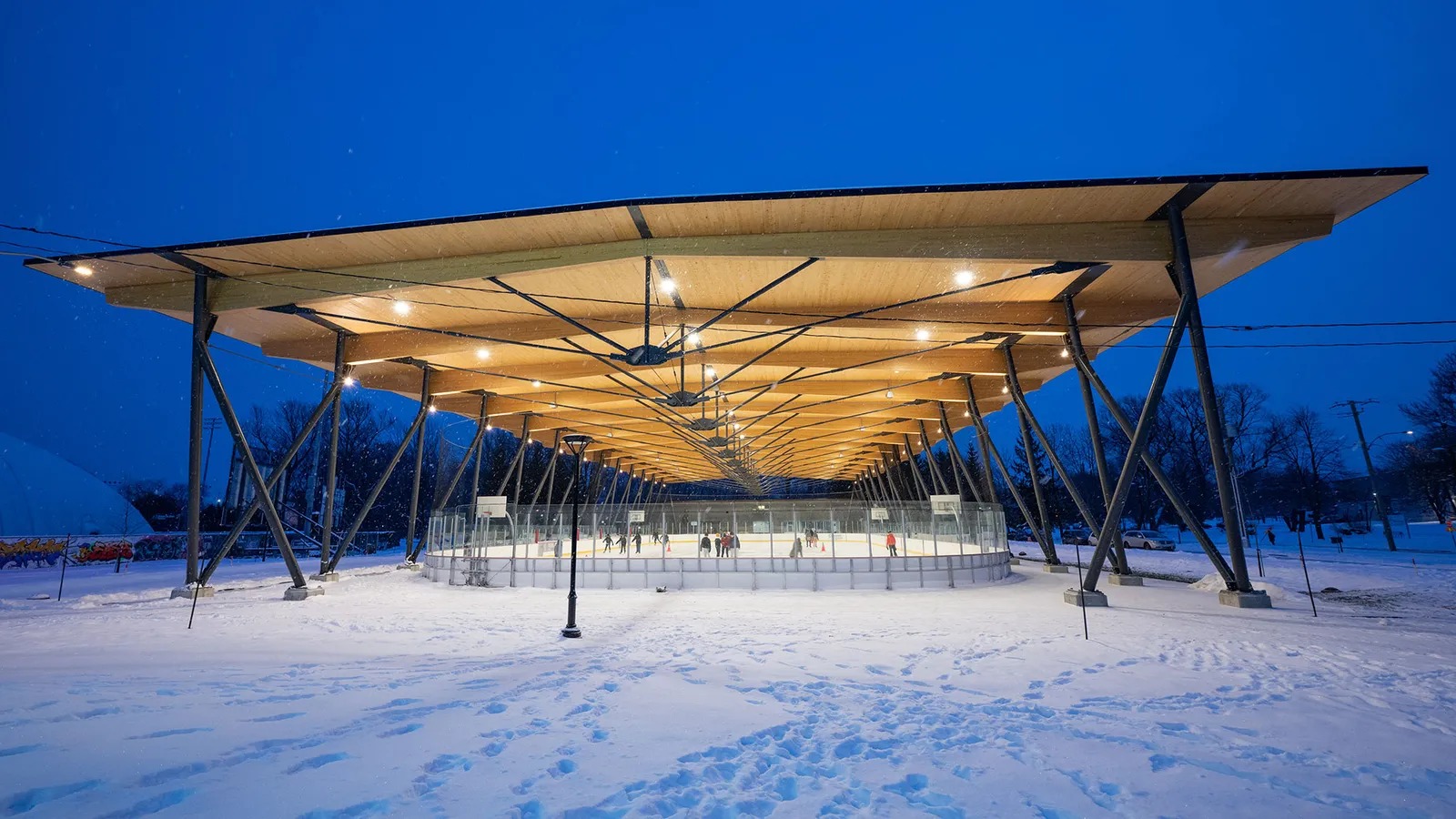 The Bleu Blanc Bouge outdoor rink in Parc Victoria, Quebec City, at dusk: a long timber canopy lit from below shelters the ice, with fresh snow in the foreground. — Patinoire Bleu Blanc BOUGE (parc Victoria)