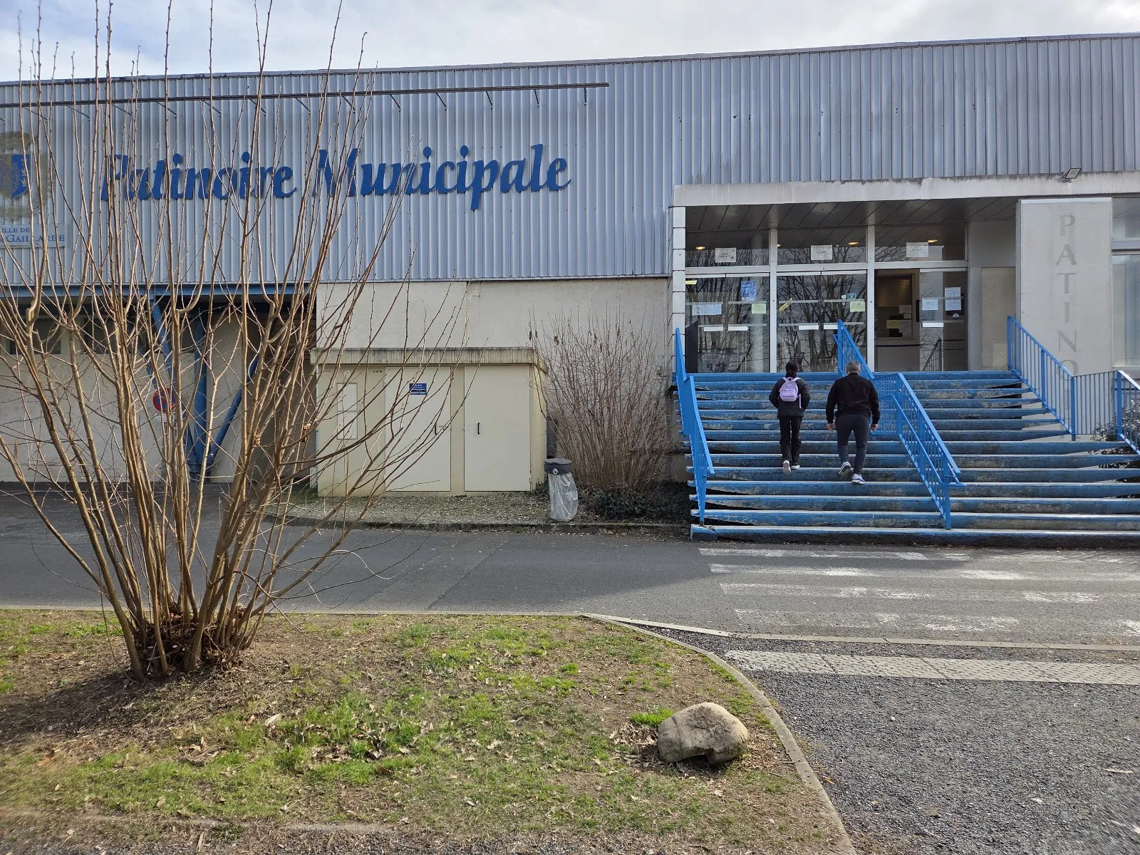 La façade grise en métal de la patinoire municipale avec ses lettres bleues, son entrée vitrée et ses marches bleues jusqu’à la porte. — Patinoire municipale de Brive
