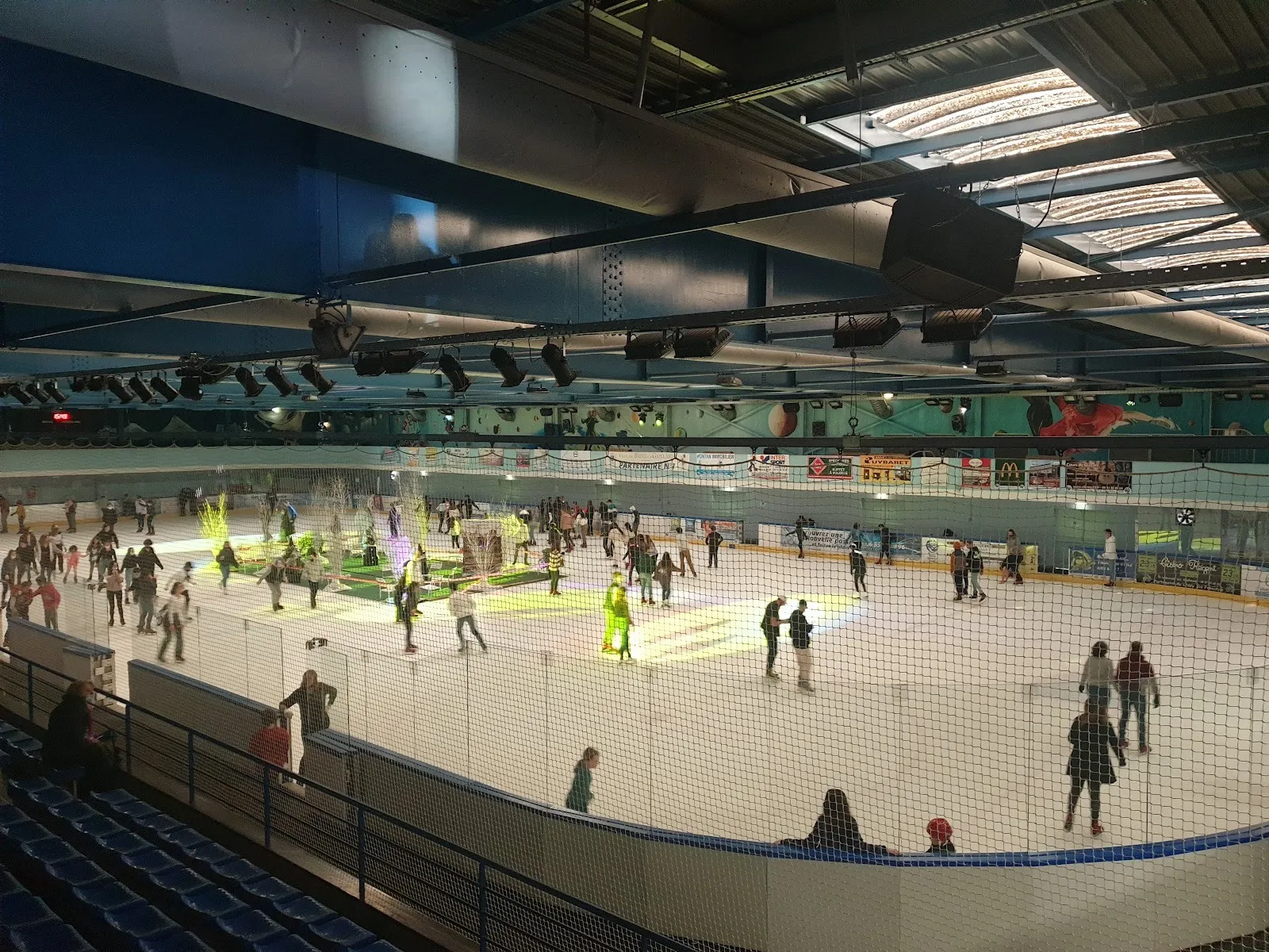 Une séance de patinage public à la patinoire municipale de Brive, des patineurs éparpillés sur la glace éclairée dans la halle, avec les gradins bleus d’un côté. — Patinoire municipale de Brive
