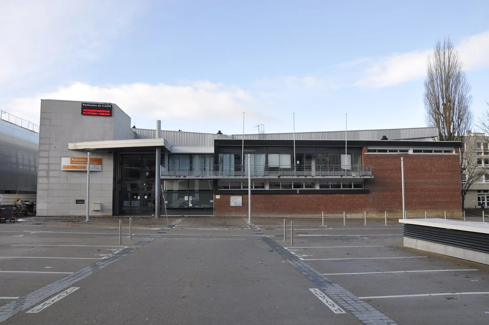 Le bâtiment de la patinoire Caen la mer vu de l’autre côté de son parking, avec un bloc d’entrée en tuiles grises, une aile en brique et l’enseigne PATINOIRE au-dessus de l’entrée — Patinoire Caen la mer
