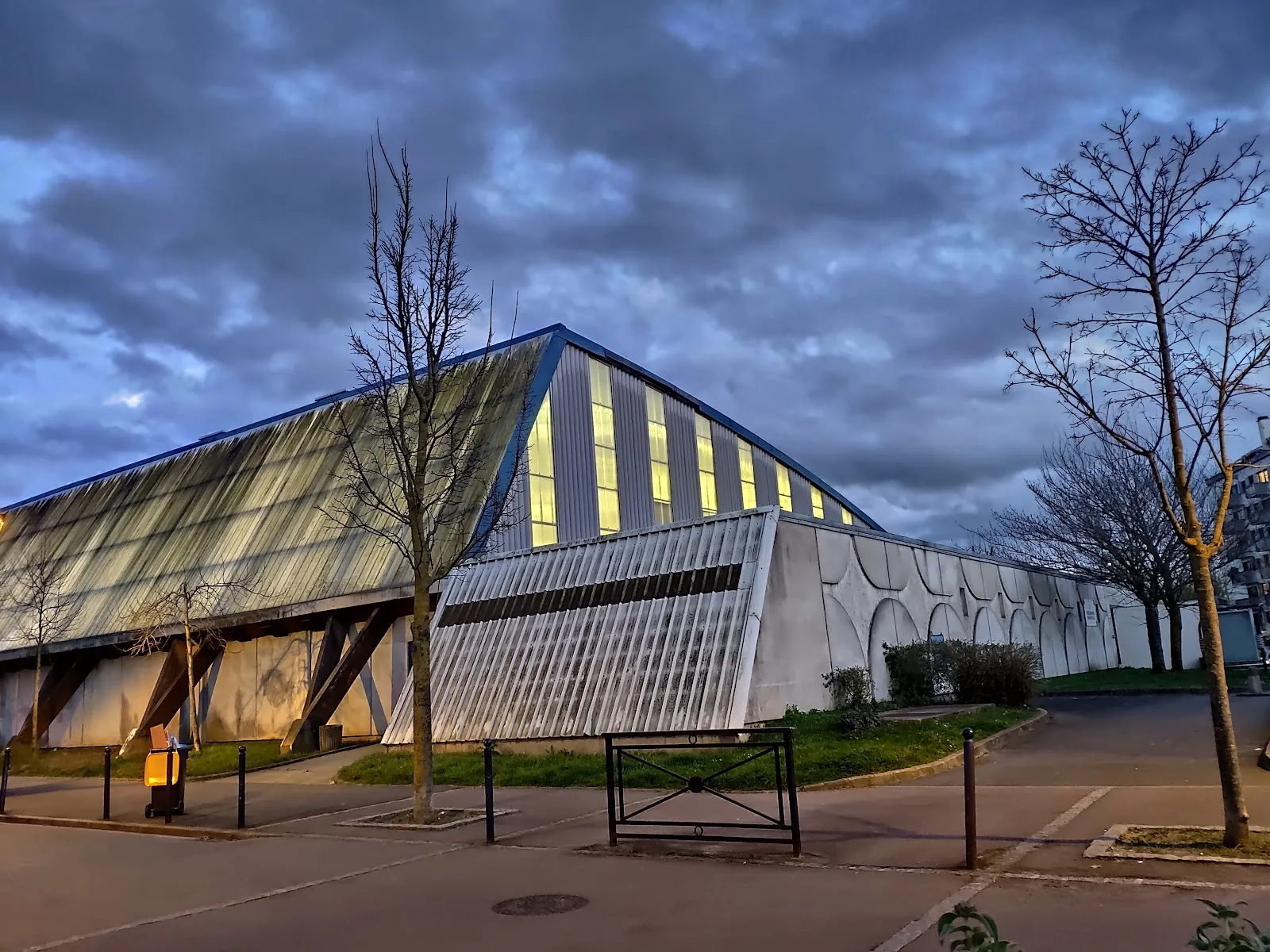 The rink building from the street: a long concrete hall with a tilted roof and a glazed gable, seen at dusk with bare trees in front. — Patinoire de Champigny-sur-Marne