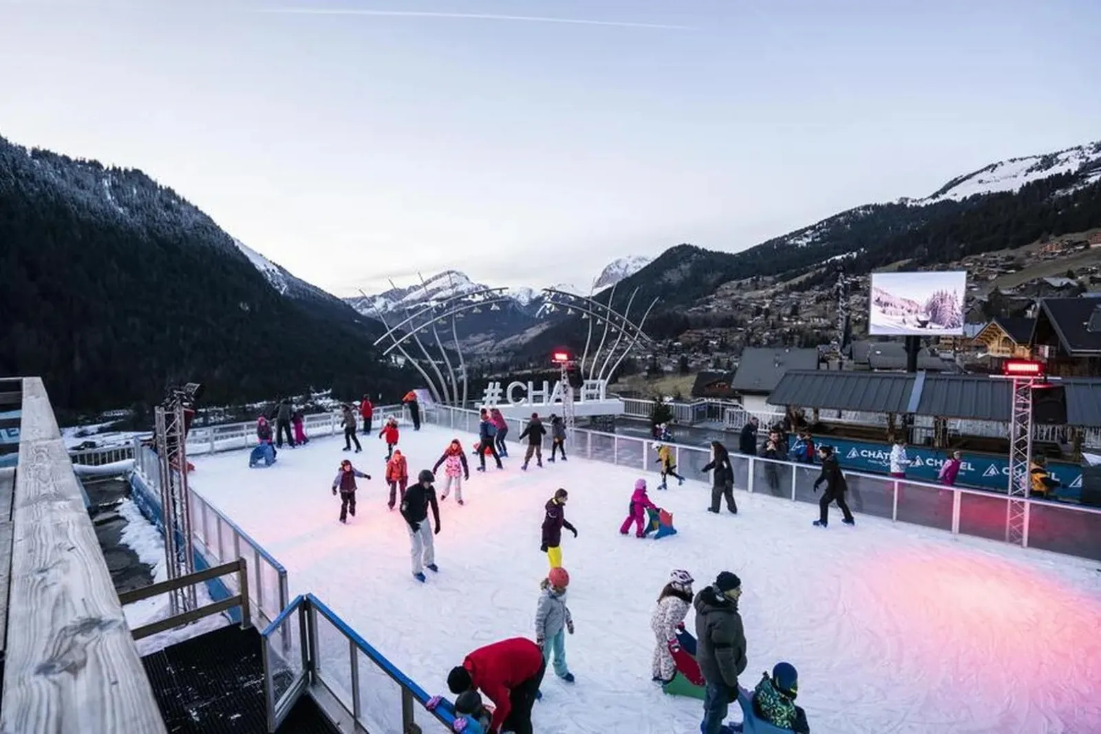 La patinoire en plein air de Châtel au milieu des montagnes enneigées, des patineurs de tous âges sur la glace avec le village et les bâtiments de la station au-delà des bandes. — Patinoire de Châtel