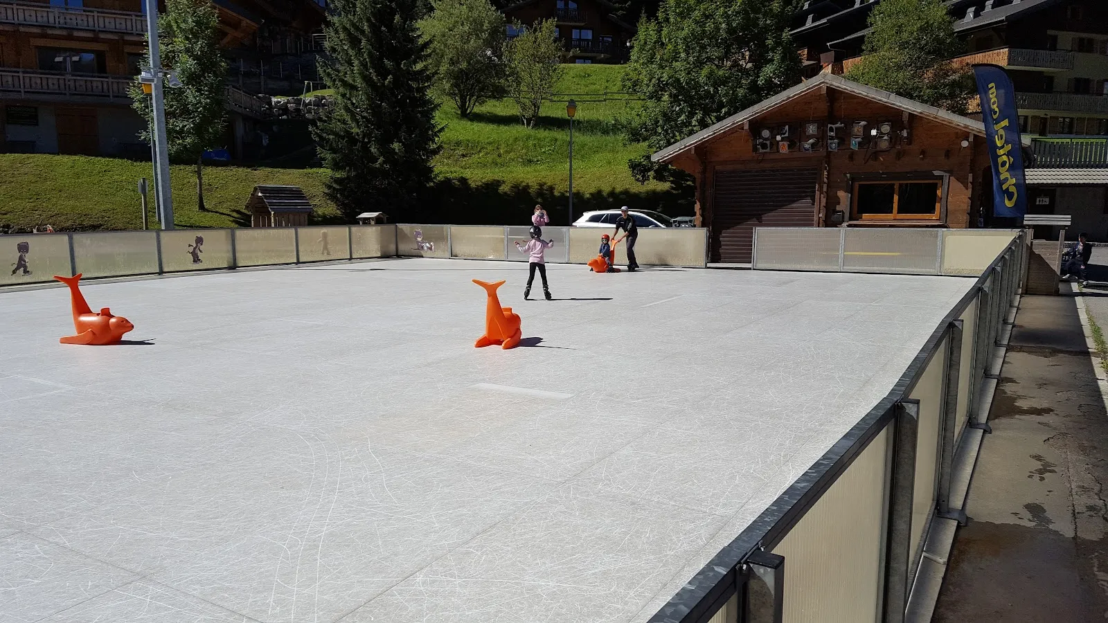 La patinoire extérieure de Châtel par une journée ensoleillée, avec ses bandes surmontées de vitres, le chalet au fond et un enfant avec un accompagnateur, loin sur la glace, parmi les aides au patinage orange en forme de phoque — Patinoire de Châtel