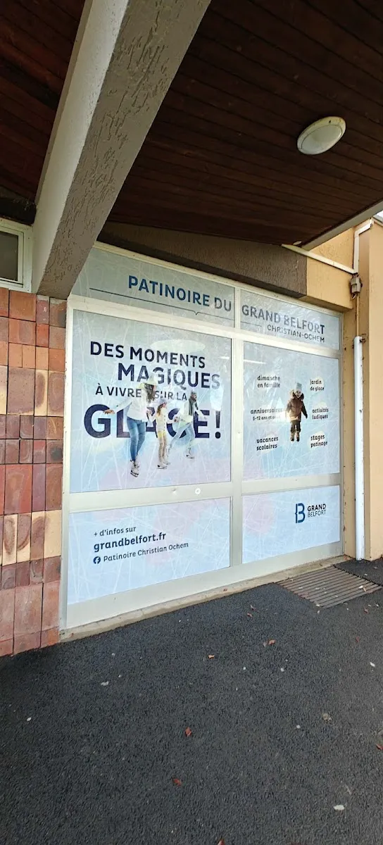 L’entrée de la patinoire du Grand Belfort Christian-Ochem sous la galerie couverte, avec le nom de la patinoire au-dessus de la façade vitrée et son affiche dans la vitrine — Patinoire Christian-Ochem
