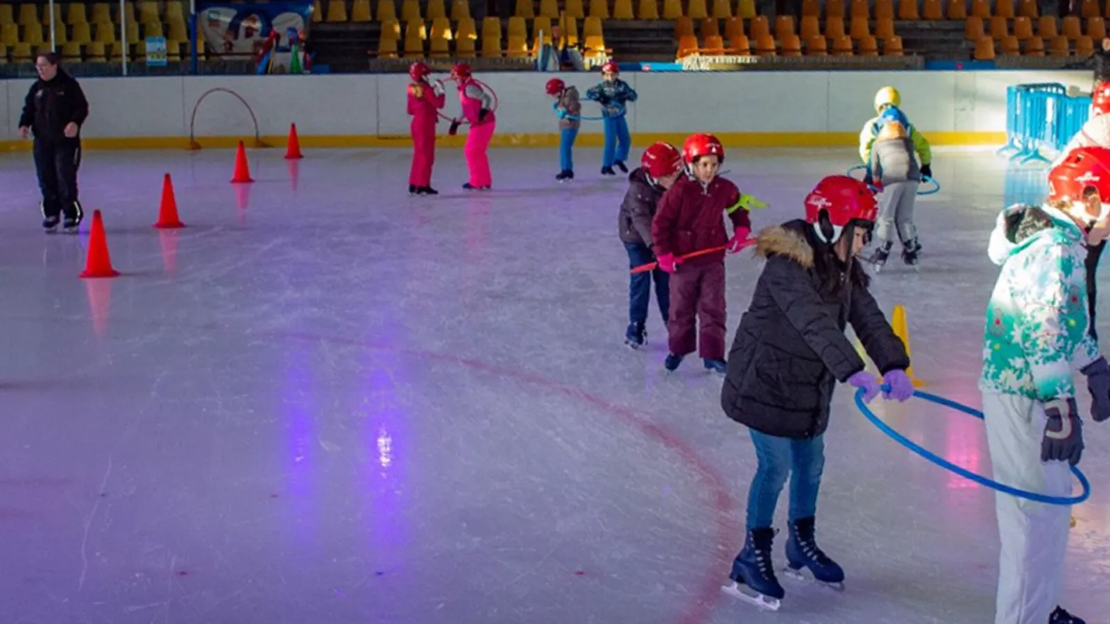 Des enfants casqués qui patinent autour de cônes et de cerceaux pendant une séance scolaire sur la glace de Bavilliers, avec les gradins derrière. — Patinoire Christian-Ochem
