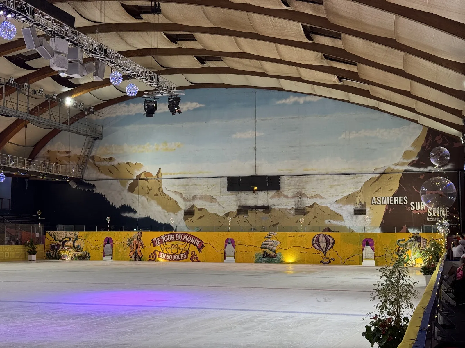 La glace vide de la patinoire des Courtilles, avec la fresque de montagne peinte derrière les bandes du fond et la voûte à nervures de bois au-dessus. — Patinoire olympique des Courtilles