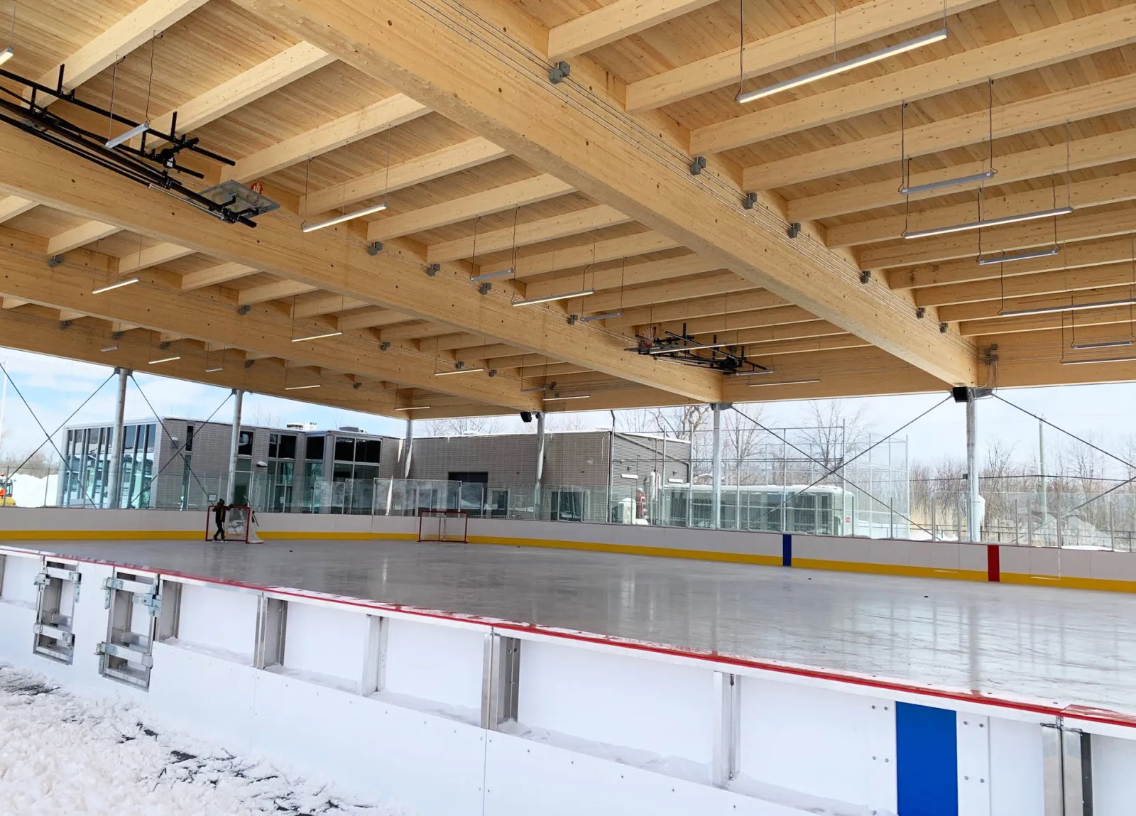 The covered rink at the Centre multifonctionnel seen from the boards, with a wide timber roof structure over the ice and glass-fronted buildings behind. — Patinoire couverte et réfrigérée du Centre multifonctionnel (Carignan)
