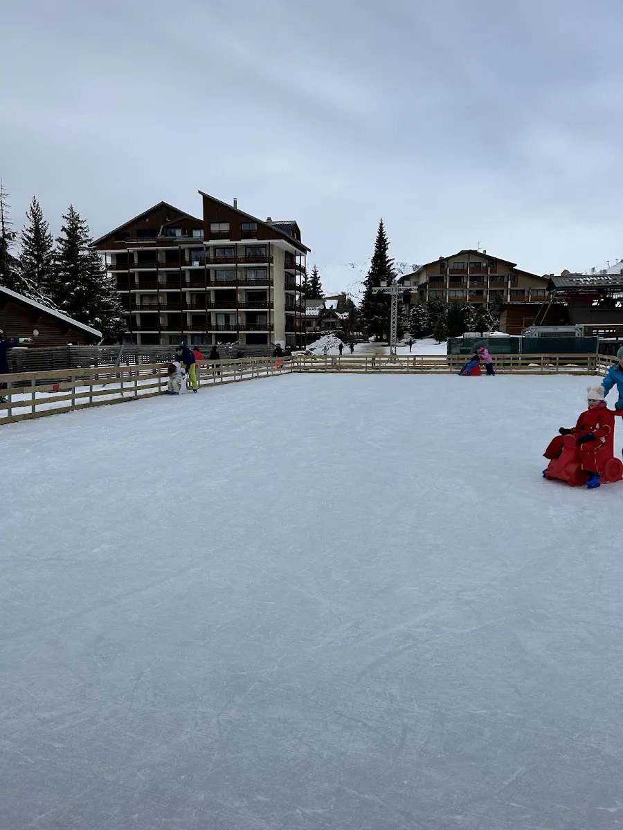 The open-air ice of the Patinoire des 2 Alpes in daylight, edged by a wooden barrier with chalet-style hotels and fir trees behind. — Patinoire des Deux Alpes