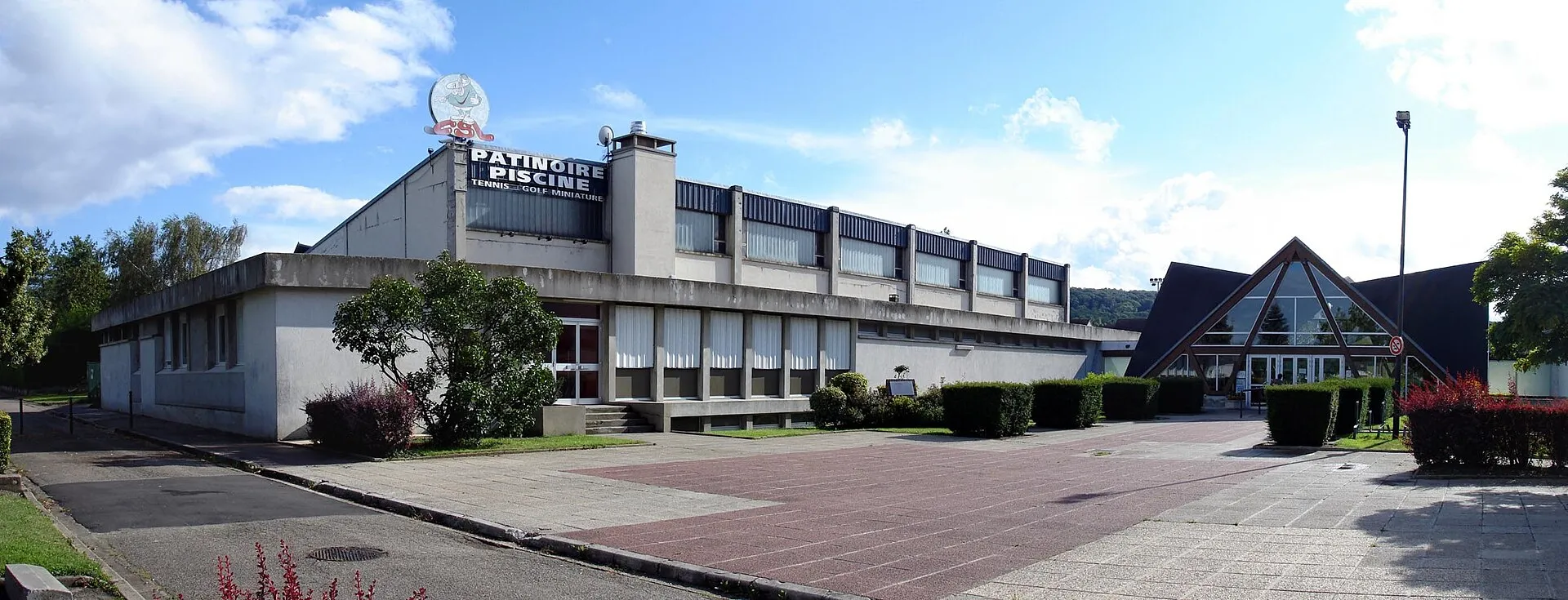 The Franconville pool and ice rink complex seen from the paved square, with the PATINOIRE PISCINE sign on the roof of the rink hall and the pitched glass entrance on the right. — Patinoire du CSL