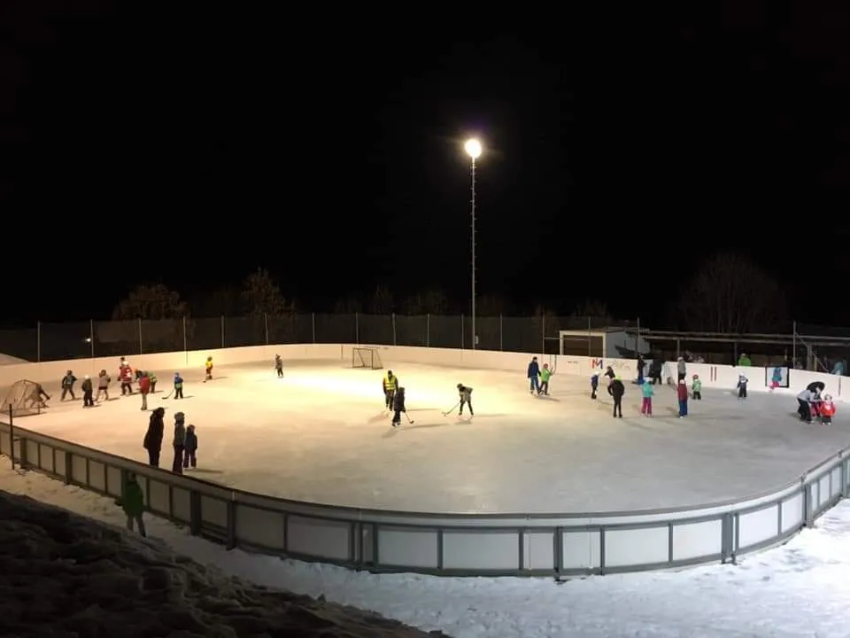 Children and adults skating and playing hockey on the floodlit outdoor rink at night, with snow banked outside the boards. — Patinoire d'Ayer