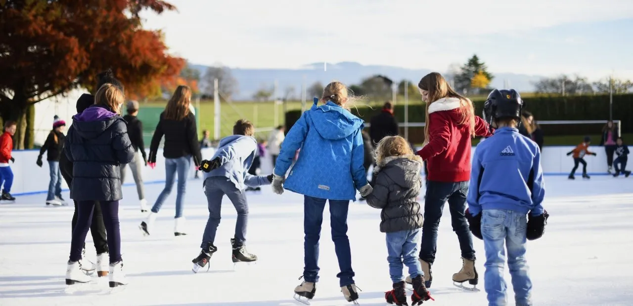 Enfants et adultes patinent, vus de dos, sur la glace blanche de la patinoire de plein air de Bernex par une journée ensoleillée, avec la bande, la pelouse et les collines au loin. — Patinoire de Bernex (Genève)