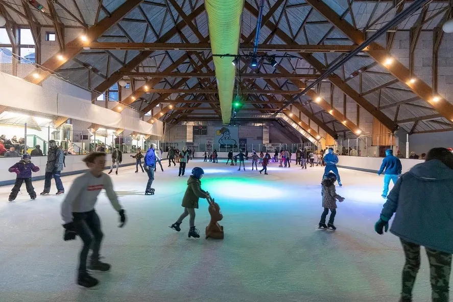 The covered Cauterets rink under its timber-trussed roof, skaters scattered across the lit ice and spectators along the boards. — Patinoire de Cauterets